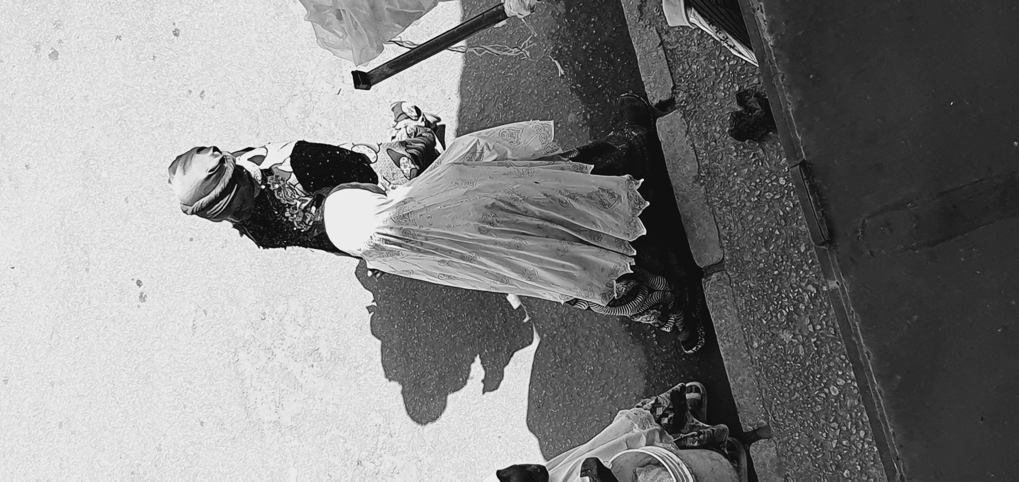 In Bukuru, Nigeria, two individuals, likely women, are depicted from a high-angle perspective on a sunlit street. The primary subject, positioned in the center and foreground, wears a light-colored, patterned veil that drapes over her body, obscuring much of her attire. Her lower legs are clad in patterned trousers or a skirt, and she wears dark footwear. To her left, another person is partially visible, also wearing dark clothing and a head covering. Both figures cast distinct shadows on the textured asphalt ground, suggesting a clear day. Scattered on the ground are various objects, including plastic bags, a white bucket, and what appears to be a broom or stick leaning against a larger plastic bag on the right. The environment appears to be a commercial or thoroughfare area, with a curb visible separating the street from a rougher, pebbled surface. The overall impression is of everyday street activity in an urban setting.