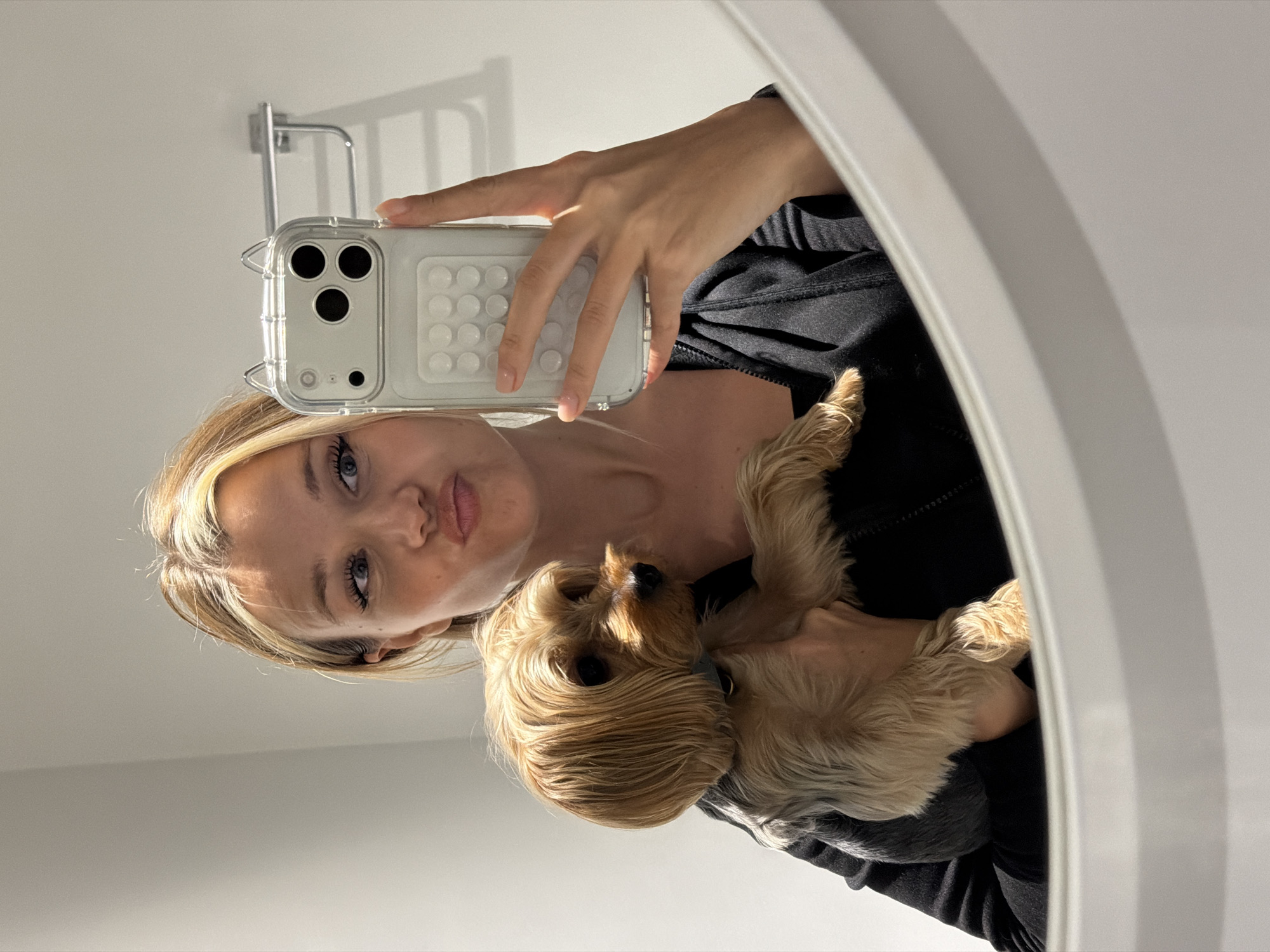 A young woman with blonde hair is taking a selfie in a mirror, holding a small, light brown Yorkshire Terrier. She is wearing a black zip-up jacket. Her mouth is puckered in a kissing pose. The dog is looking to its right. The background appears to be a bathroom with white walls and a towel rack. The reflection in the mirror shows the woman's face, upper body, and the dog. The image does not provide any information about the city of Thep Krasatti, Thailand.