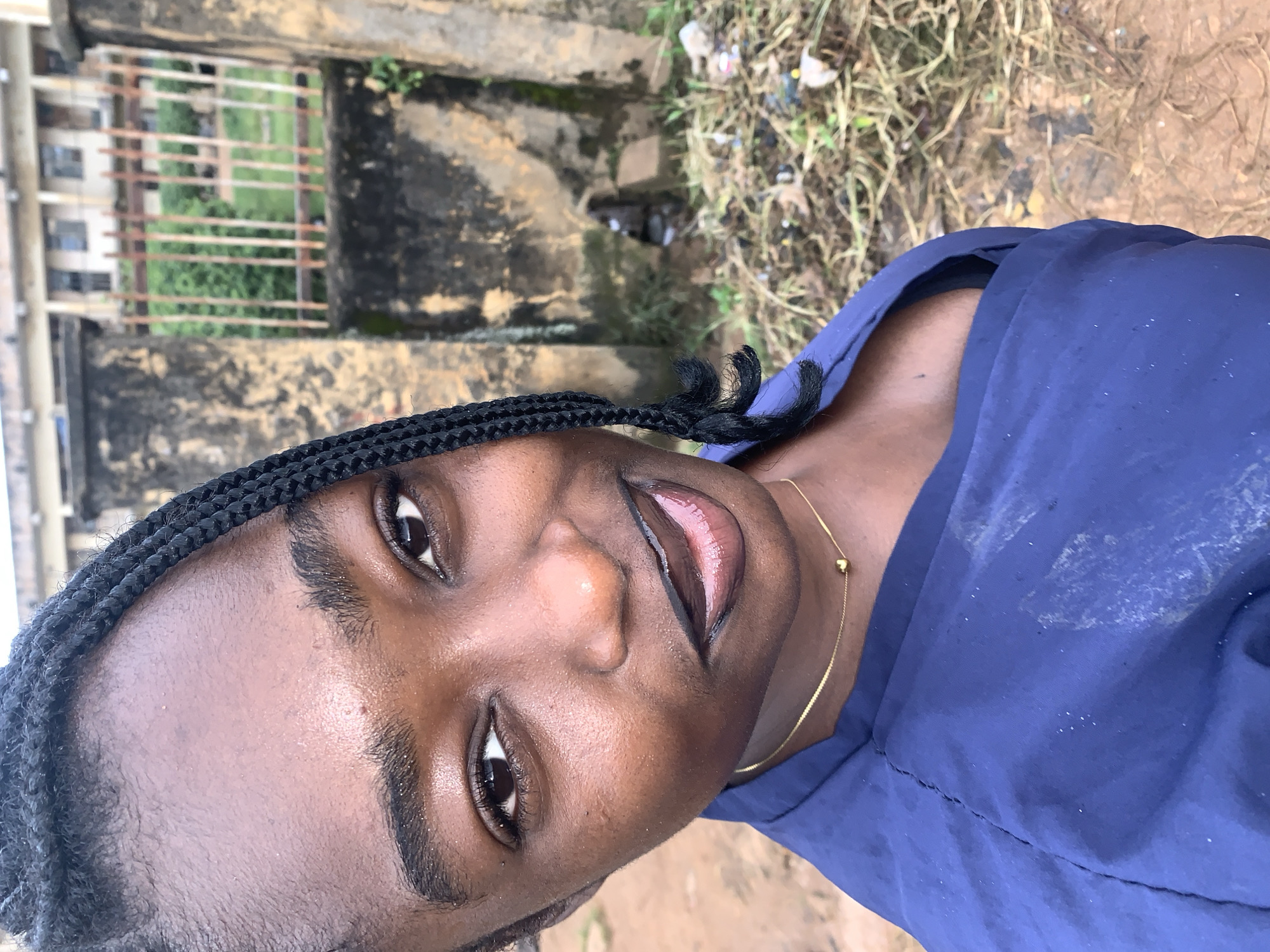 A close-up, eye-level, outdoor medium shot shows a young Black woman with dark skin and braided hair, facing the camera. Her hair is styled into dark, thin braids that are pulled back from her face, with a few strands framing her forehead. She is wearing a dark blue v-neck top and a thin gold chain with a small heart pendant. Her makeup includes dark eyeliner and a dark, glossy lipstick. She has a slight smile, with her lips slightly parted. Her skin appears to be dewy.
The background is out of focus. To the left of the woman, a blurry, textured concrete wall is visible, with some mossy patches. Beyond the wall, there is a rusty metal fence, behind which green foliage and a light-colored building with multiple windows can be seen. To the right of the woman, the ground appears to be covered in dry grass, dirt, and scattered debris, including some plastic wrappers. The overall impression is that of an outdoor environment within a populated area, possibly a residential or institutional setting, given the building in the background. The ground suggests an unkempt or neglected area.