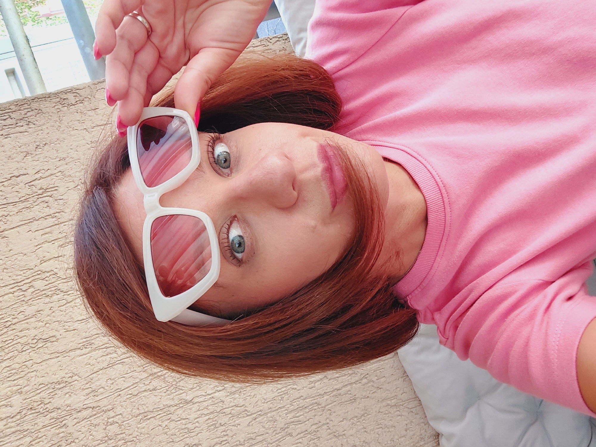 A woman with shoulder-length reddish-brown hair wears large, white-framed sunglasses with pink lenses and a bright pink t-shirt. She is holding the sunglasses up with her right hand, revealing her left eye, which is blue-green. Her lips are slightly pursed. The background appears to be an outdoor setting with a textured beige wall and a white cushioned surface, possibly a couch or lounger. The image is a close-up selfie, suggesting the woman is taking the photo herself. The scene does not provide any specific visual cues to identify a location within Sofia, Bulgaria.