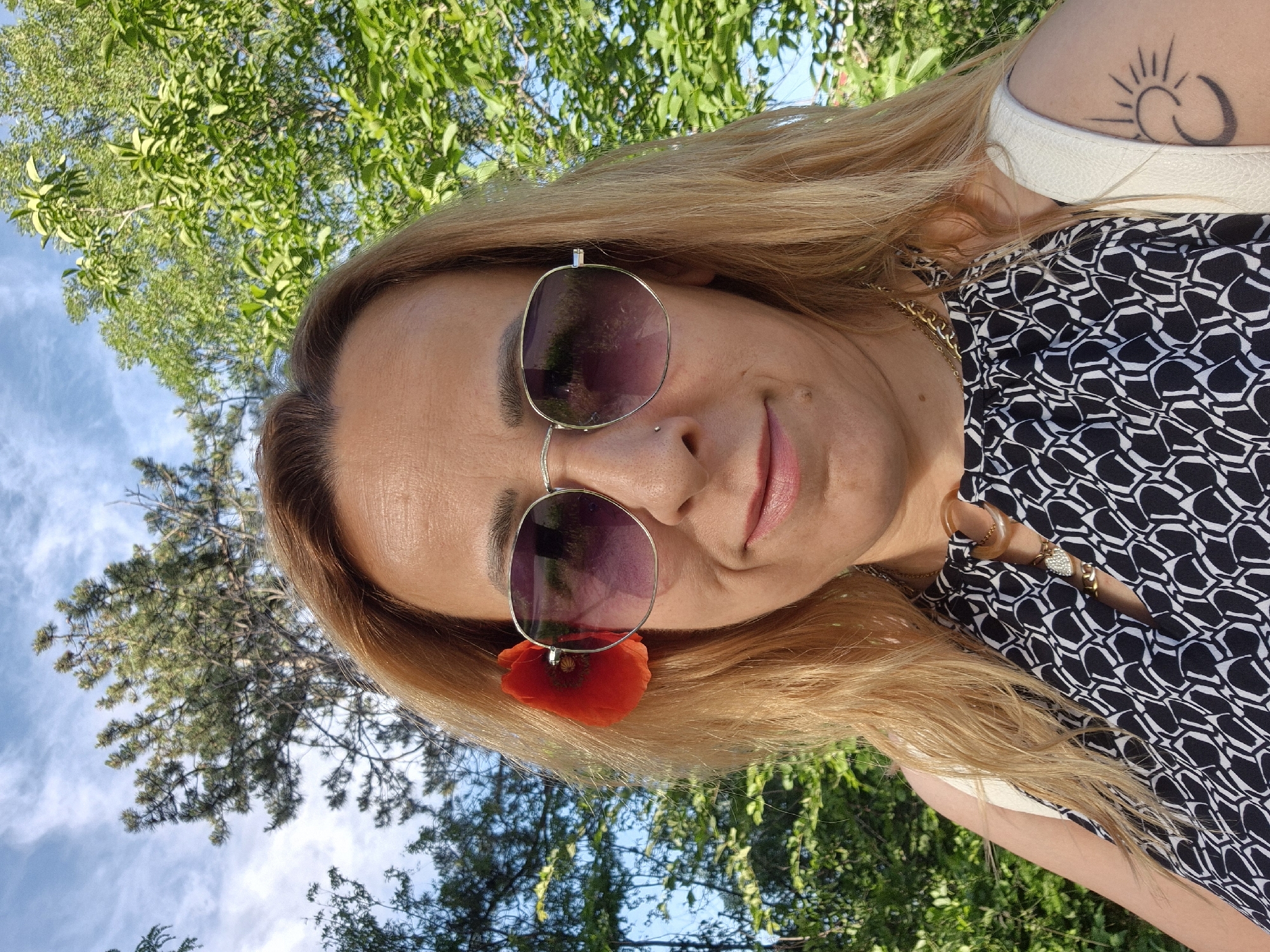 A woman with blonde hair is wearing sunglasses with a red flower tucked into one lens. She has a nose piercing and a tattoo of a sun-like figure on her shoulder. She is wearing a black and white patterned shirt. The background includes trees and a blue sky with clouds. The location is unknown.