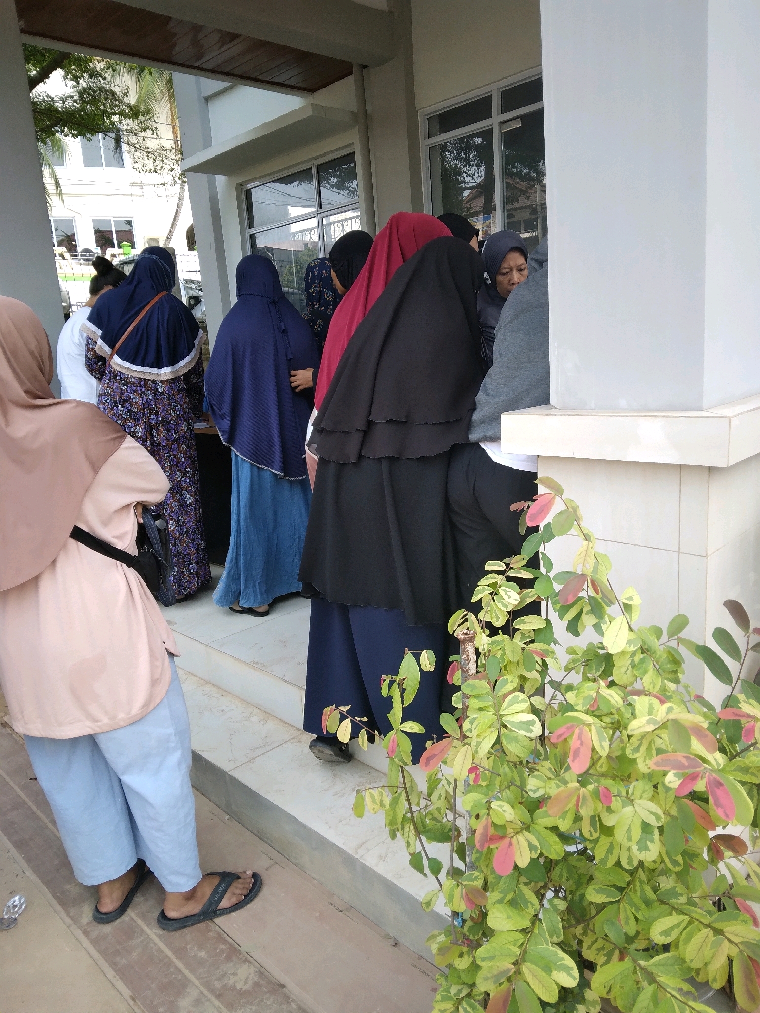 The image depicts a group of individuals, predominantly women wearing hijabs, standing on a tiled outdoor walkway in front of a building. The building appears to have a white facade with windows and a covered overhang supported by columns. One woman in the foreground is wearing a light pink top and light blue pants, with black flip-flops. Behind her, several other women are visible, dressed in various colored hijabs and modest clothing, including a woman in a dark blue hijab and patterned dress, and another in a blue hijab and long blue skirt. Some individuals appear to be engaged in conversation. A lush green plant with variegated leaves, featuring touches of pink and yellow, is positioned in the lower right corner of the frame, partially obscuring the building's base. The overall setting suggests a public or institutional area, possibly near an entrance. The architectural style and vegetation are consistent with urban environments in Batam, Indonesia.