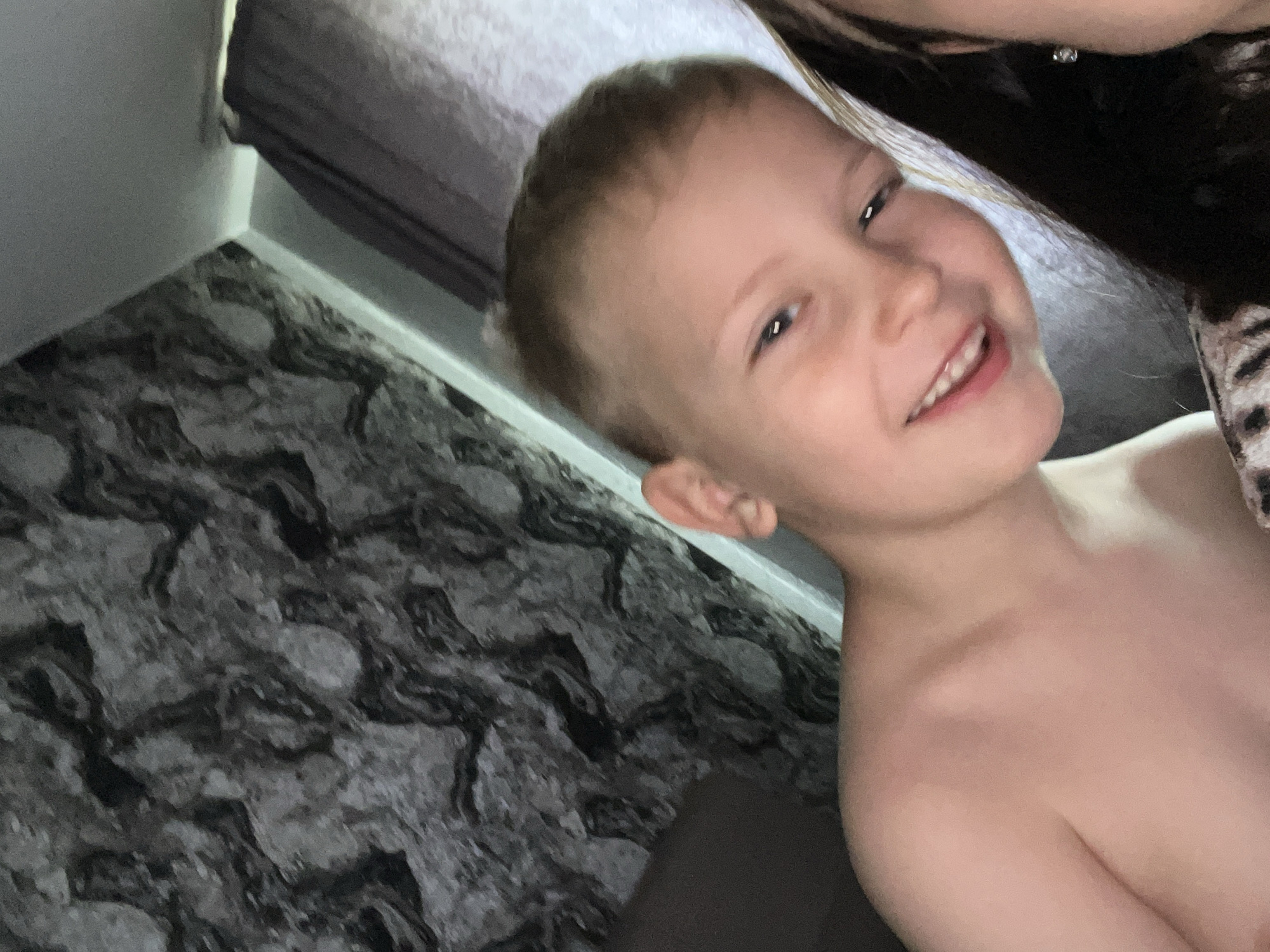 The image shows a young child with short brown hair, fair skin, and a smiling expression. The child's upper torso is visible and appears to be unclothed. A person with dark hair is partially visible in the upper right corner of the frame. The background includes a gray patterned wall and a gray cushion. The location of the image is Unknown, Unknown.