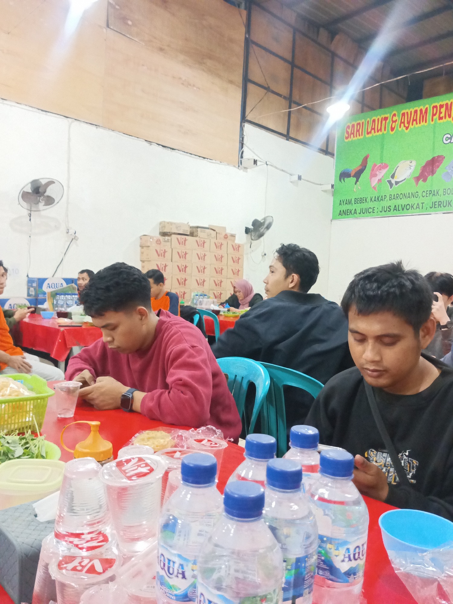 The image depicts a casual dining scene, likely at a restaurant or food stall, set within an indoor environment with fluorescent lighting. The color palette is dominated by red, as seen on the tablecloths, and muted tones of wood and white on the walls and ceiling.
In the foreground, several plastic bottles of "Aqua" brand bottled water are arranged on a red surface, some with blue caps. Several clear plastic cups, some with red branding that is difficult to decipher, are also present, stacked and scattered. A green plastic basket containing some sort of leafy greens or vegetables is visible on the left. A yellow condiment dispenser with a red lid is also on the table.
Several individuals are seated at the tables, engaged in eating or socializing. A young man in a maroon sweatshirt is in the middle ground, looking down at his phone, with a clear plastic cup in front of him. To his right, another young man in a black sweatshirt is also looking down, his hands partially visible. Further back, other patrons are seen at tables, their faces mostly obscured. A woman wearing a pink hijab is visible in the background.
Above the dining area, a large green banner with text in Indonesian is displayed, featuring images of fish and a rooster. The text appears to advertise seafood and chicken dishes, as well as juices. The overall impression is a busy, informal dining establishment.
While the specific location in Makassar, Indonesia, is not explicitly identified in the image, the presence of "Aqua" bottled water, the Indonesian text on the banner, and the general ambiance are consistent with dining establishments found in such urban Indonesian settings.