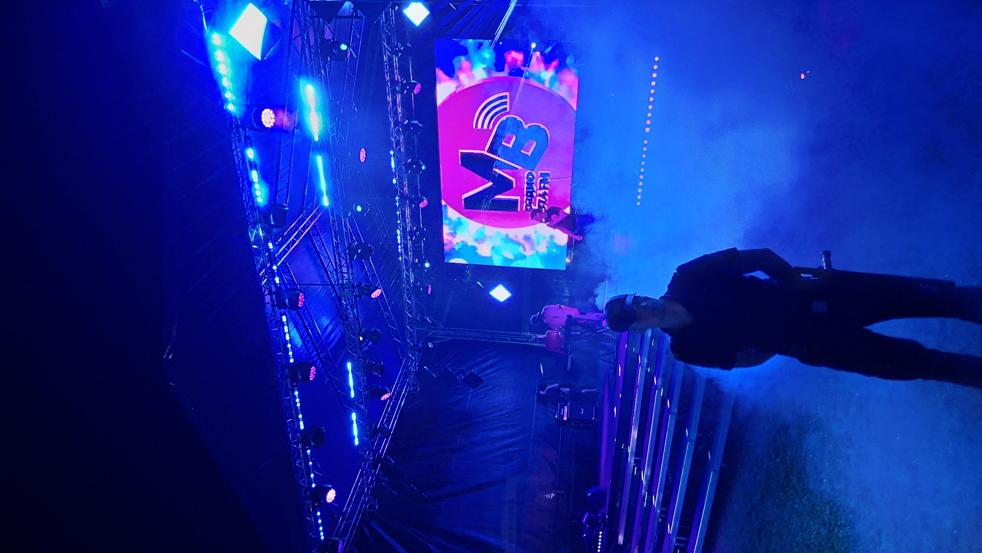 The image depicts an outdoor stage setup illuminated by vibrant blue and pink lights. In the foreground, a person wearing a dark polo shirt and cap stands in a haze of blue smoke. Behind them, a stage is set up with seating on the left and a large screen displaying a logo with the letters "MB" and some text in a Cyrillic script, possibly indicating a radio station. Several individuals are visible on the stage, appearing to be performers or crew members. The overall atmosphere suggests a nighttime event, possibly a concert or festival, taking place in an open area. While the image itself does not provide specific location data within Myadzyel, Belarus, such a venue could be a park, town square, or dedicated event space within the city.