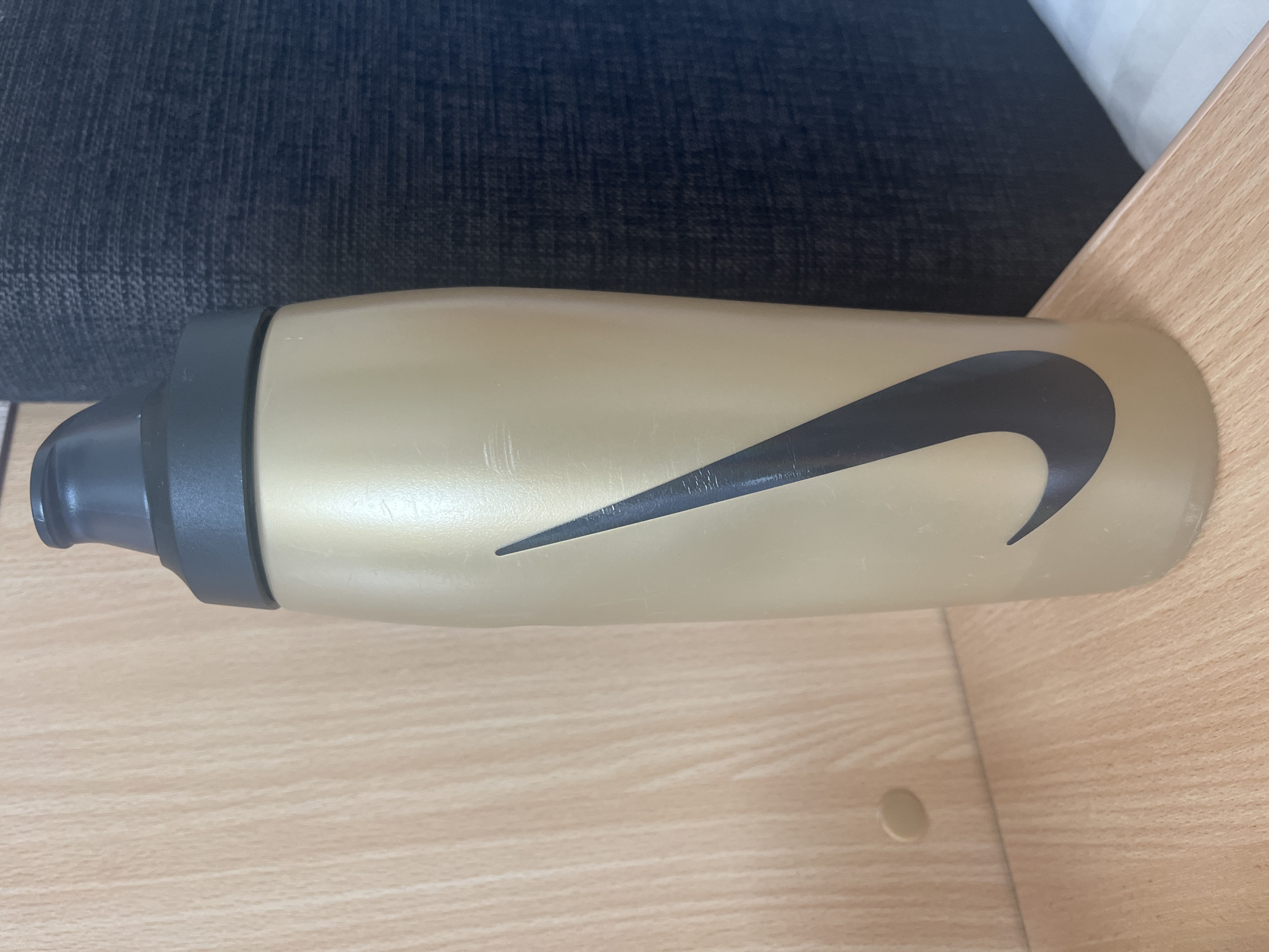 A gold-colored Nike water bottle with a black Nike swoosh logo is standing upright on a light-colored wooden surface. The bottle has a black cap and appears to be made of plastic. Behind the bottle, a dark textured fabric is visible, which could be part of a sofa or chair. The background on the left is a light-colored wooden wall with visible grain. There is a small, round, light-colored object on the wooden wall to the left of the bottle, possibly a knot in the wood or a small decorative element. The lighting suggests an indoor environment. No people are present. The scene is indoors and does not provide specific details about the city of Toila, Estonia.