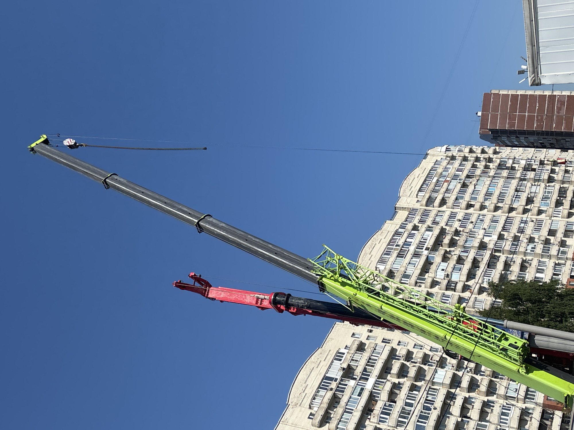 The image captures a vibrant, high-angle view of two large cranes against a clear blue sky, set within an urban landscape. In the foreground, a prominent black and grey crane boom extends diagonally upwards from the lower left towards the upper right. Attached to its tip, a hook block with a chain is visible, extending further into the sky. Below this crane, a bright green crane boom runs parallel, also extending upwards and diagonally. This green crane features a red jib or attachment at its base, closer to the viewer.
In the background, a large, multi-story building with a light-colored facade is visible. It has numerous rectangular windows and balconies arranged in a repetitive, modular pattern, suggesting a residential or office complex with a distinctive architectural style. Parts of its curved upper sections are also visible. Behind and to the right of this building, the upper section of another, darker building with a brown or reddish-brown top is partially seen. A small portion of green foliage, likely a tree, is visible between the green crane and the main building.
The scene depicts a construction or heavy lifting environment. The clear blue sky and bright illumination indicate it is daytime with sunny weather. The photograph is taken from a low vantage point, looking up at the cranes and buildings, emphasizing their height against the sky. No discernible text is visible in the image.