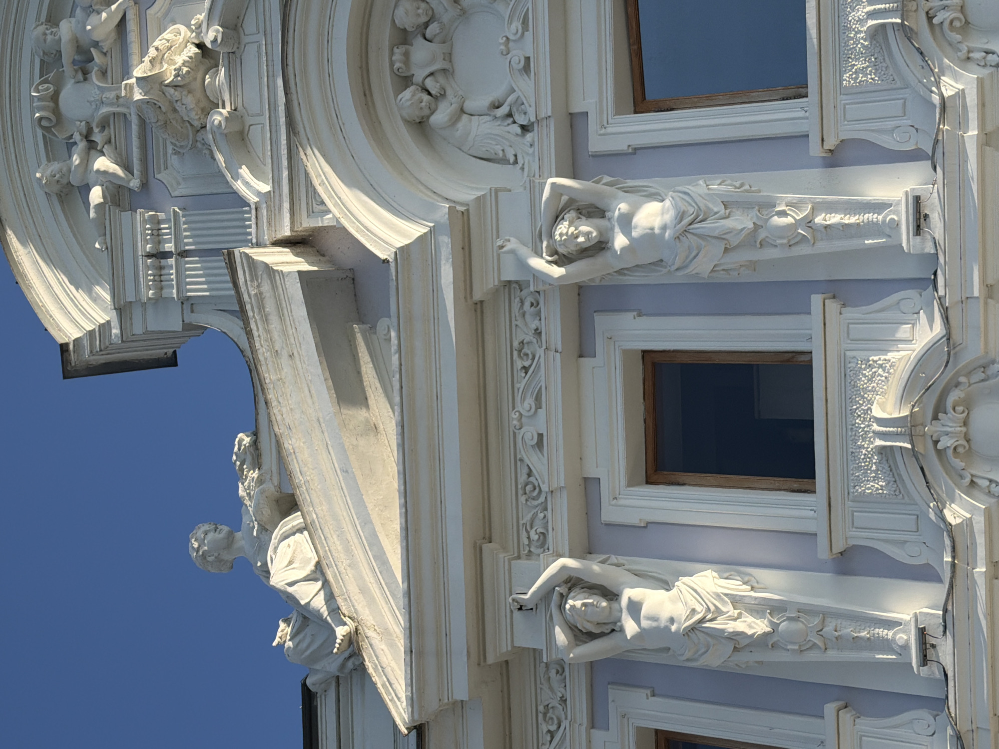 This is a low-angle, exterior, close-up, full-frame shot of an ornate, white-stone building facade against a clear, bright blue sky. The architecture features elaborate sculptural decorations, including statues of women and cherubs, as well as intricate relief work. In the upper section of the image, a reclining female statue is visible on a sloped ledge. Below this, two standing female statues, depicted as caryatids, support the upper architectural elements. Their arms are raised, and they are draped in flowing garments. In the upper right corner, a relief of two cherubs interacting with a decorative element can be seen. The building has light purple walls visible between the white decorative sections. The windows on the facade are dark and recessed. The lighting suggests a sunny day, with strong shadows highlighting the three-dimensional details of the sculptures and architectural features. The image is a detail of the facade of the House of Melnikov, located in Nizhny Novgorod, Russia.
