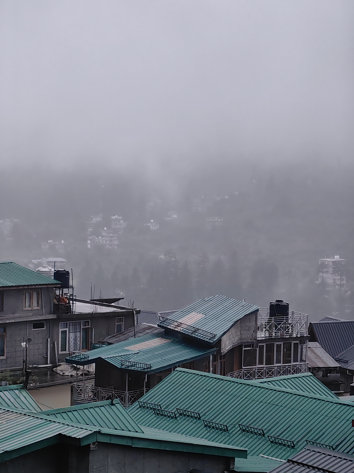 This is a high-angle, full shot of a hazy mountain town in Vashisht, India. The foreground is dominated by rows of buildings with green, corrugated metal roofs. The roofs are angled and vary in size, with some displaying decorative black metal grates or elements.
In the mid-ground, more buildings are visible, also with predominantly green roofs. One building features a water tank on its rooftop. Beyond this, the town continues to spread across a hilly terrain, but visibility is severely reduced by a thick, gray fog. The fog obscures the details of distant structures, creating a soft, indistinct background where only the outlines of buildings and trees can be faintly discerned. The overall impression is one of a damp, overcast day, possibly during or after rain, given the wet appearance of some surfaces. No people or specific actions are discernible in the image.