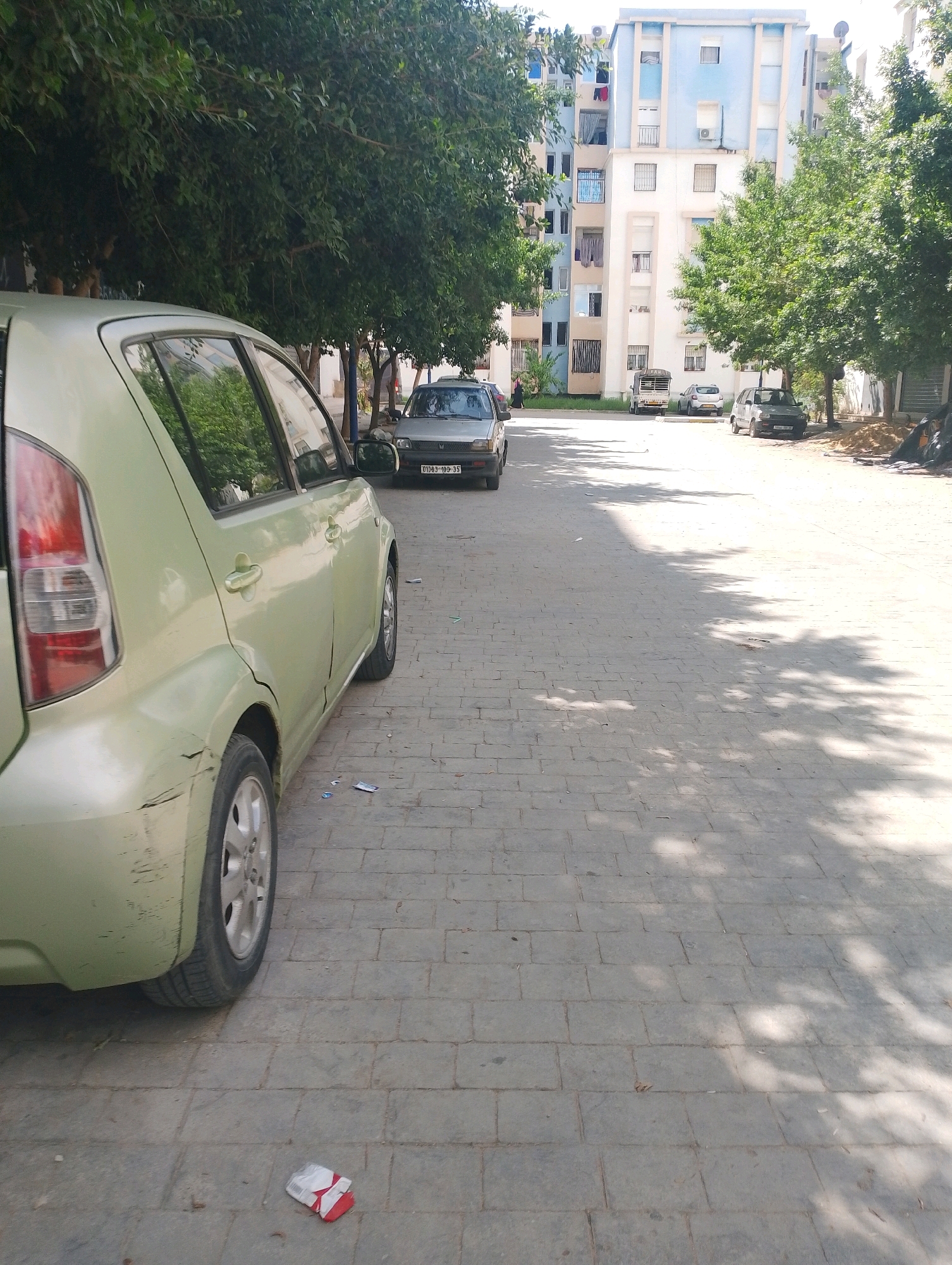 The image captures a sun-drenched street scene in Bordj Menaiel, Algeria. In the foreground, a light green, four-door hatchback car is parked, its rear section visible, displaying damage to the bumper and bodywork. The car is positioned on a paved sidewalk composed of rectangular gray bricks. Further down the street, several other vehicles are parked, including a silver hatchback and a darker colored sedan with visible license plates. In the background, a tall, multi-story apartment building with blue and white facade dominates the skyline. Trees with lush green foliage line the street, casting dappled shadows on the pavement. The overall impression is a typical residential area in a North African city on a clear, bright day.