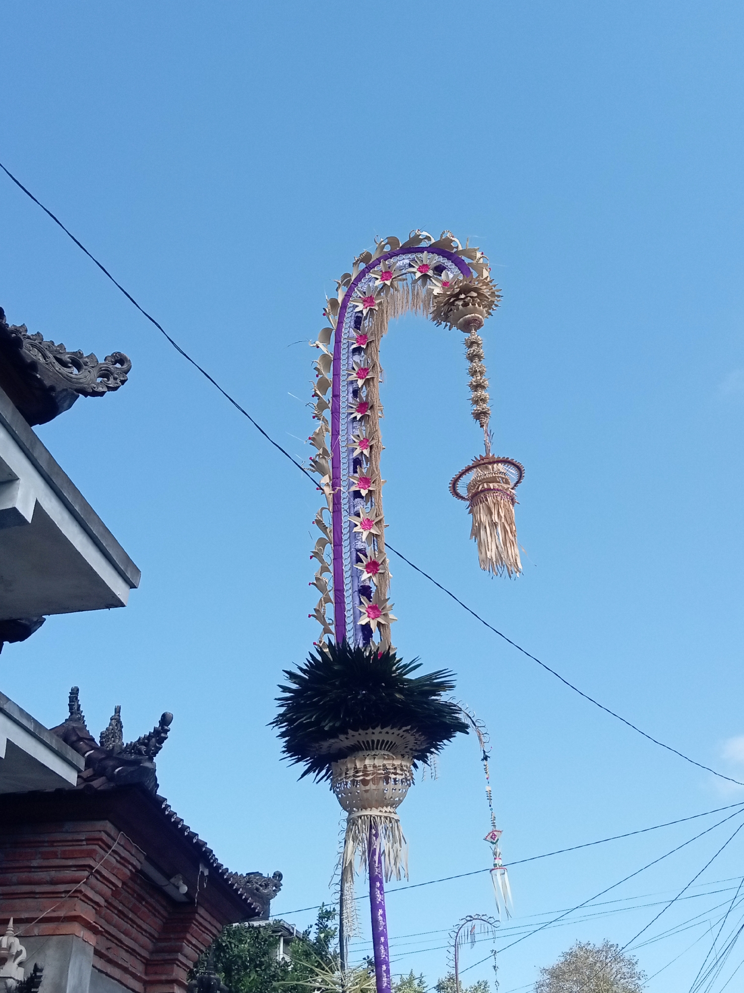 A tall, ornate, curved banner pole, likely a penjor, stands against a clear blue sky. The banner pole is decorated with woven palm leaves, purple fabric, and pink floral accents. A decorative pendant hangs from the top of the pole, fashioned from dried palm leaves. The base of the penjor is adorned with a large, dark green leafy structure. To the left, a portion of a traditional Balinese building is visible, featuring intricate stone carvings on its dark tile roof and a red brick wall. Electrical wires crisscross the sky in the background. The scene appears to be outdoors, under bright daylight.