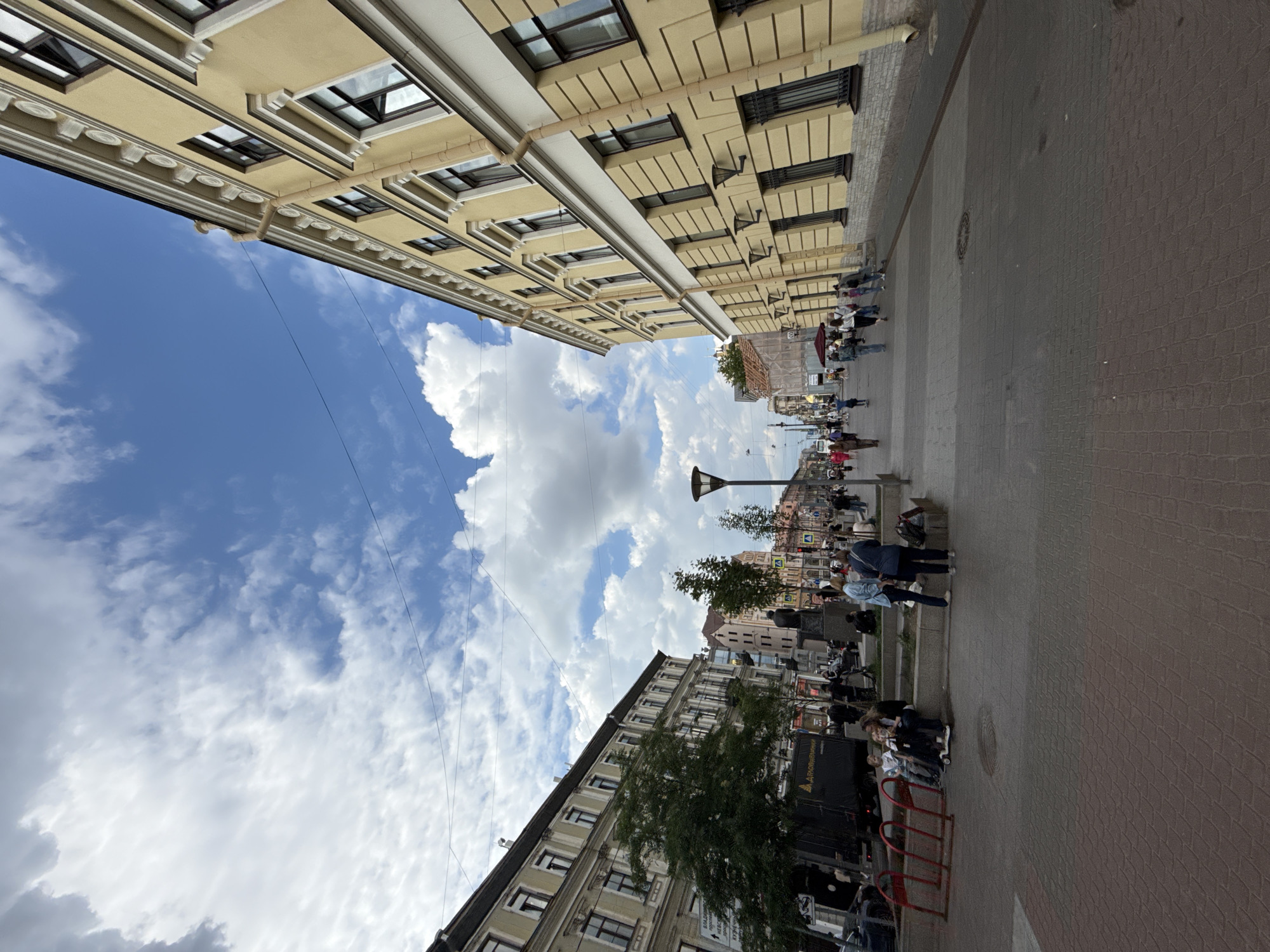 The image captures a vibrant urban street scene, looking down a thoroughfare lined with buildings under a bright, cloudy sky. The scene is oriented as if looking from a slightly low angle, with tall buildings flanking both sides and the sky dominating the upper part.
Numerous pedestrians are visible on the paved sidewalks and street, engaged in walking, standing, and congregating. Some individuals are gathered around outdoor tables, suggesting cafes or restaurants.
The buildings on the left are multi-story, featuring light yellow or beige facades with classical architectural details such as cornices, decorative window frames, and relief work. Buildings on the right, further down the street, are also multi-story with similar architectural styles.
The sky above is clear blue with scattered, fluffy white cumulus clouds. Multiple thin, dark overhead wires crisscross the sky between the buildings. A modern-style dark streetlight stands on the right, along with a few small trees offering greenery. The ground is paved with interlocking bricks or pavers in shades of light brown and reddish-brown.
In the mid-ground on the right, a dark truck is parked. White text on its side is partially visible, reading "ДАРКОВЩИНА" and "АРЕНДА ГРУЗОВИКОВ" (meaning "Truck Rental"). In the foreground on the right, red metal bicycle racks are present. The time of day appears to be daytime, with bright ambient light and fair weather.