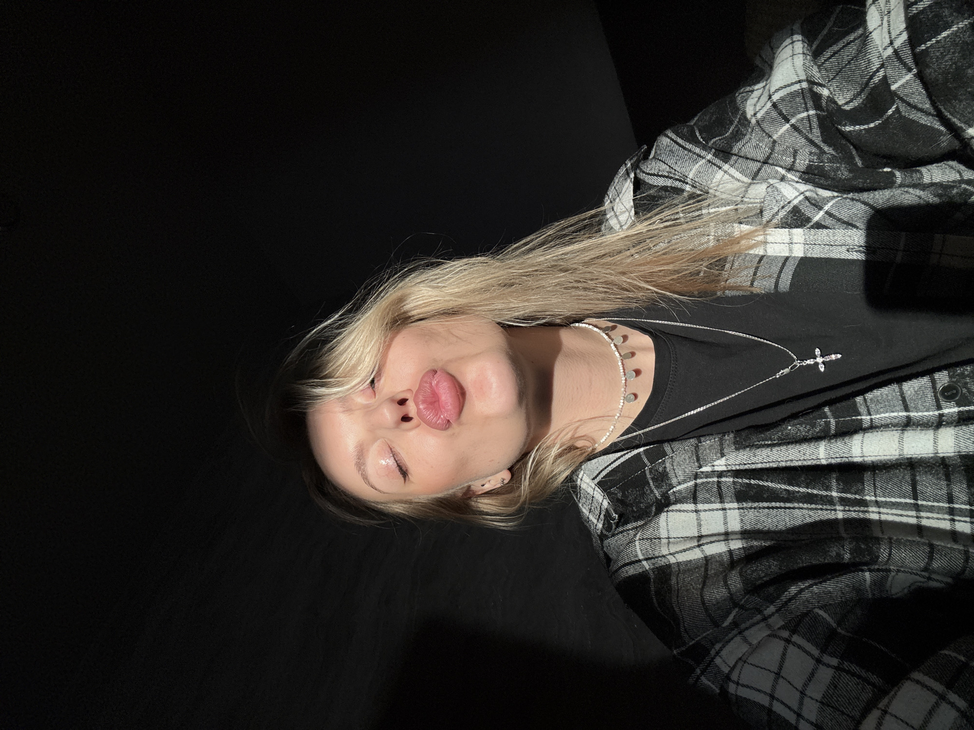 A fair-skinned individual with blonde hair is seen in a photograph. The person has their eyes closed and lips puckered. They are wearing a black t-shirt, a silver necklace with a cross pendant, and a black and white plaid flannel shirt. The background is black. The location, Krasnoyarsk, Russia, cannot be confirmed from the image alone.