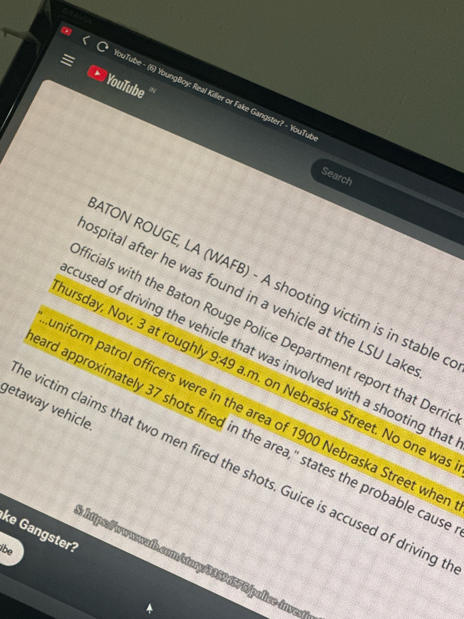 The image shows a close-up of a laptop screen displaying a news article about a shooting incident. The article, from Baton Rouge, LA (WAFB), reports on a shooting victim who was found in a vehicle at the LSU Lakes. Officials with the Baton Rouge Police Department are investigating the incident, which occurred on Thursday, November 3rd, at approximately 9:49 a.m. on Nebraska Street. The article highlights that "uniform patrol officers were in the area of 1900 Nebraska Street when the shots were fired." The victim claims that two men fired approximately 37 shots. The report also mentions that a suspect, Guice, is accused of driving the getaway vehicle. The top of the screen shows a YouTube page with the title "(6) YoungBoy: Real Killer or Fake Gangster? - YouTube".
