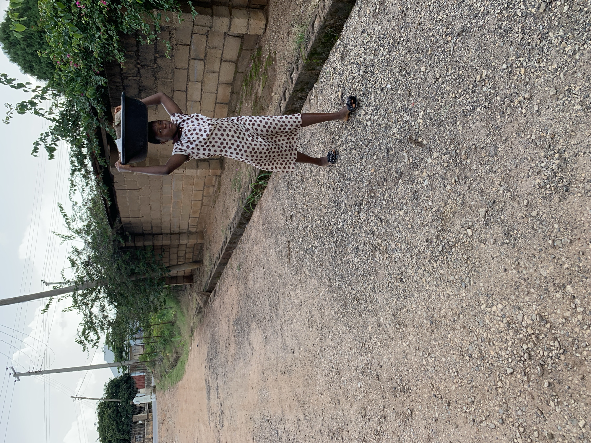 A young woman is walking on a gravel road in Jos, Nigeria, carrying a large black basin full of laundry on her head. She is wearing a light-colored dress with brown polka dots and dark flip-flops. The road is lined with a low brick wall and overgrown vegetation. In the background, there are power lines and trees. The sky is overcast.