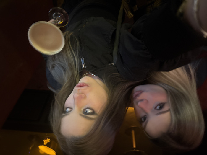 Two young women are captured in a selfie-style photograph, likely taken indoors at night in a dimly lit bar or restaurant. The image features the women positioned close together, both with long, light brown hair.
The woman on the left, looking directly at the camera with a relaxed and slightly playful expression, has prominent blue-grey eyes and slightly parted lips. She is dressed in a dark jacket, and a silver-toned chain is visible around her neck. Her arm is raised, holding a martini glass filled with a light brown, creamy beverage, possibly a cocktail like an espresso or chocolate martini, positioned above her head.
The second woman, on the right side of the frame, is partially obscured by the first woman's head and hair. She also has long, light brown hair and is looking slightly away from the camera, with a more subdued expression. She is also wearing dark clothing.
The background is dark and out of focus, with blurry warm lights and reflections, suggesting an intimate and social atmosphere typical of an evening outing. There is no visible text within the image. The overall impression is one of friends enjoying a night out.