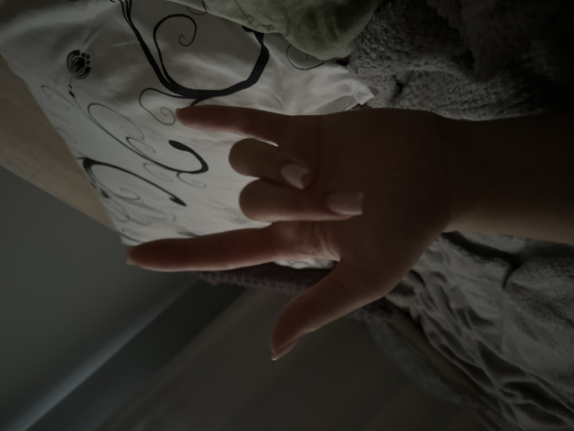 A close-up, low-angle, medium shot shows a person's left hand, with light pink-tipped nails, making the "rock on" gesture. The hand is in the foreground and partially obscuring a bed in the background. The bed has a white pillow with black floral patterns, a grey textured blanket, and a green quilted item visible. The lighting is dim, with shadows cast across the scene, suggesting an indoor environment at night or in low light conditions. The location is Unknown, Unknown.