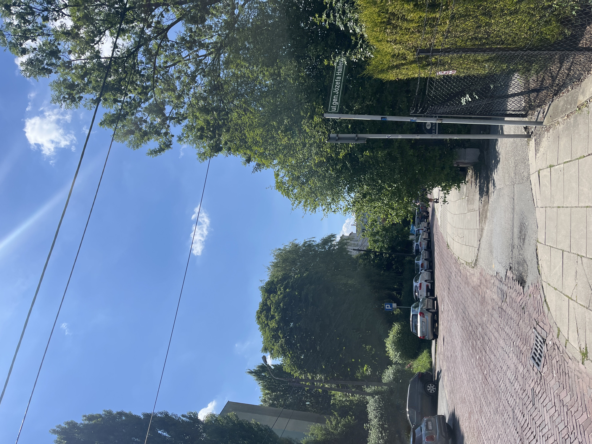 The image depicts a street scene on a sunny day, likely in the late morning or early afternoon given the bright sunlight and clear blue sky. The perspective is from a low angle, looking up towards the sky and across a street lined with parked cars. The street itself is paved with reddish-brown bricks, with a sidewalk made of grey concrete alongside it. Lush green trees with full foliage line the street and cast shadows on the ground, suggesting a warm season like spring or summer.
A street sign is visible, reading "ul. gen. Józefa Hallera". There are several cars parked along the brick-paved street, most of them silver or grey. One car closer to the foreground is dark grey. Power lines run diagonally across the sky, and a few scattered white clouds drift by. A metal fence with some greenery behind it is also present. The overall impression is one of a quiet, residential street on a pleasant day. There are no people prominently visible in the frame. The focus is on the environment, with the cars and streetscape as the main subjects. The sunlight creates a bright and cheerful atmosphere.