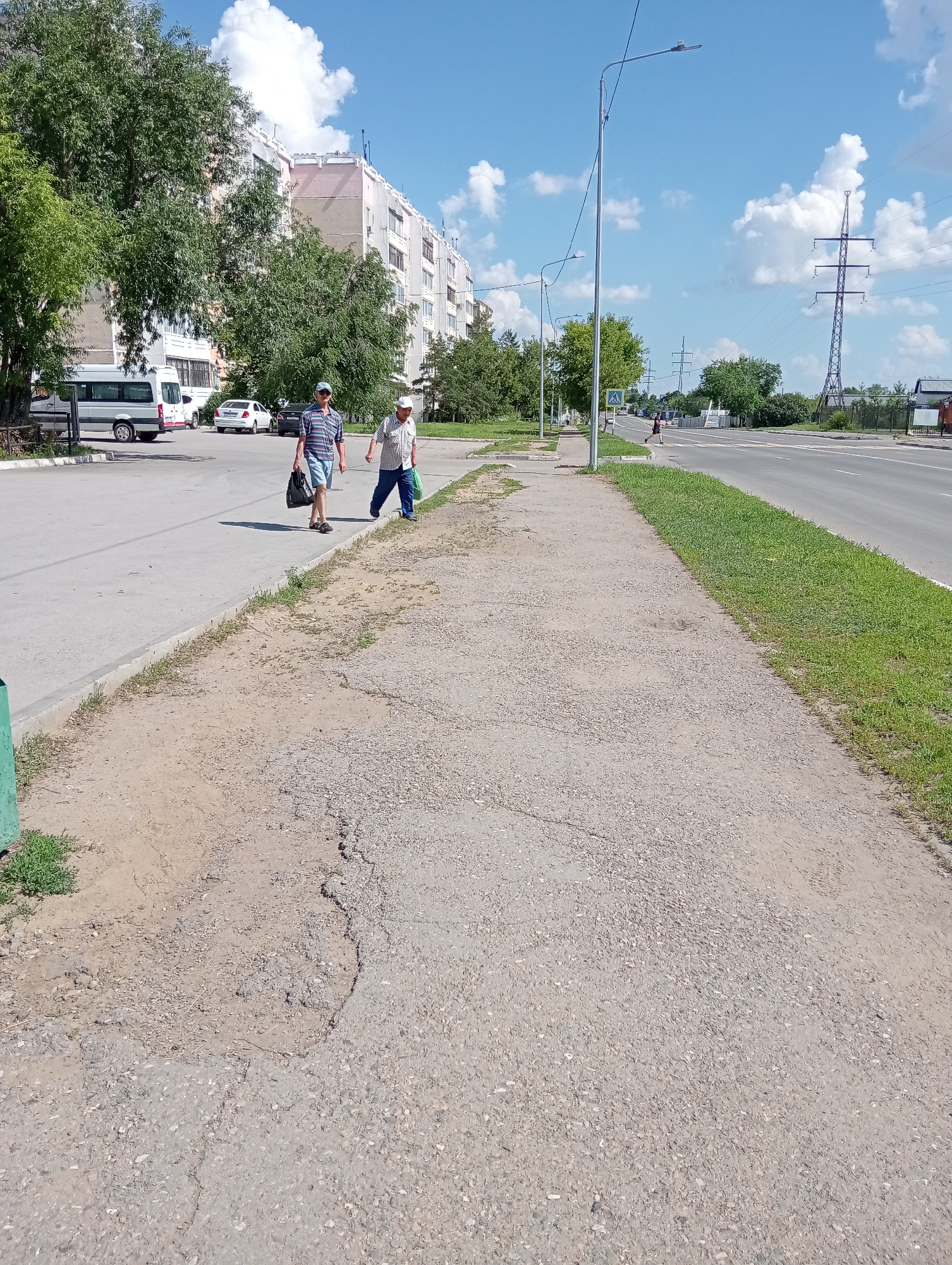 The image depicts a daytime scene on a city street. Two men are walking on the sidewalk. The man on the left, wearing a striped shirt and shorts, carries a black bag. The man on the right, in a white shirt and a cap, holds a blue bag. Behind them, a white van and several cars are parked along the street. Further down the road, there is a pedestrian crossing and a traffic light. Tall trees line the sidewalk, and a multi-story building is visible in the background. The sky is blue with scattered white clouds, and a power line tower stands in the distance. The sidewalk appears to be paved and somewhat worn.