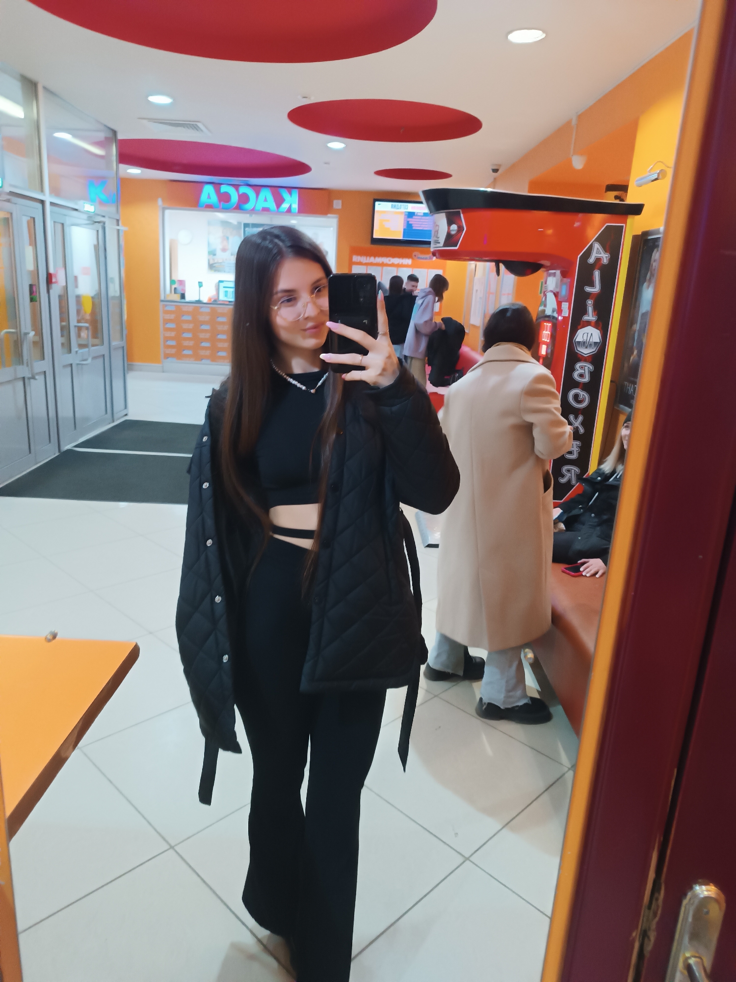 A young woman is taking a selfie in what appears to be a public indoor space, possibly a shopping mall or entertainment center. She is wearing black clothing including a top, pants, and a quilted jacket. The environment has an orange and red color scheme with tiled floors. In the background, there are other people, a seating area, and what looks like a boxing game machine. There is a sign with the word "KASSA" on it, likely indicating a ticket or payment area. The location is in an urban area, in the city of Unknown, Unknown.