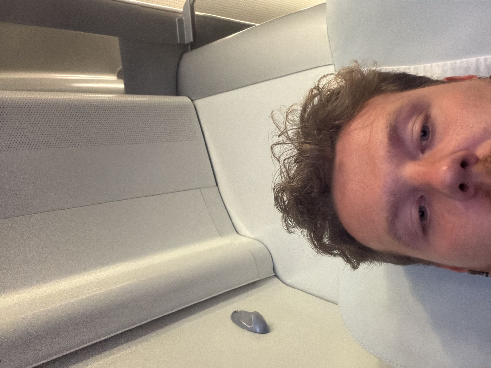 A man with curly brown hair and light skin is taking a selfie. He is seated in a white, modern-looking passenger compartment, possibly a train or bus. A small gray object, possibly an earbud, is resting on the white surface near him. The interior has a smooth, minimalist design. The location appears to be inside a vehicle, but the city of Milton Keynes cannot be determined solely from this image.