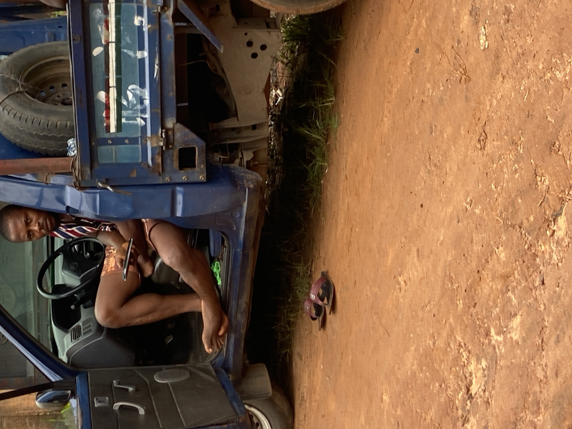 A man sits in the driver's seat of a blue utility truck with the door open, on a dirt road in Nkpor, Nigeria. He is wearing a red, white, and blue striped shirt and shorts, and is holding a smartphone. His left leg is extended out of the truck, with his bare foot resting on the ground. A pair of maroon flip-flops are on the ground near the truck's door. The truck has a spare tire mounted on the back. The background shows dry, dusty earth and some sparse green grass.