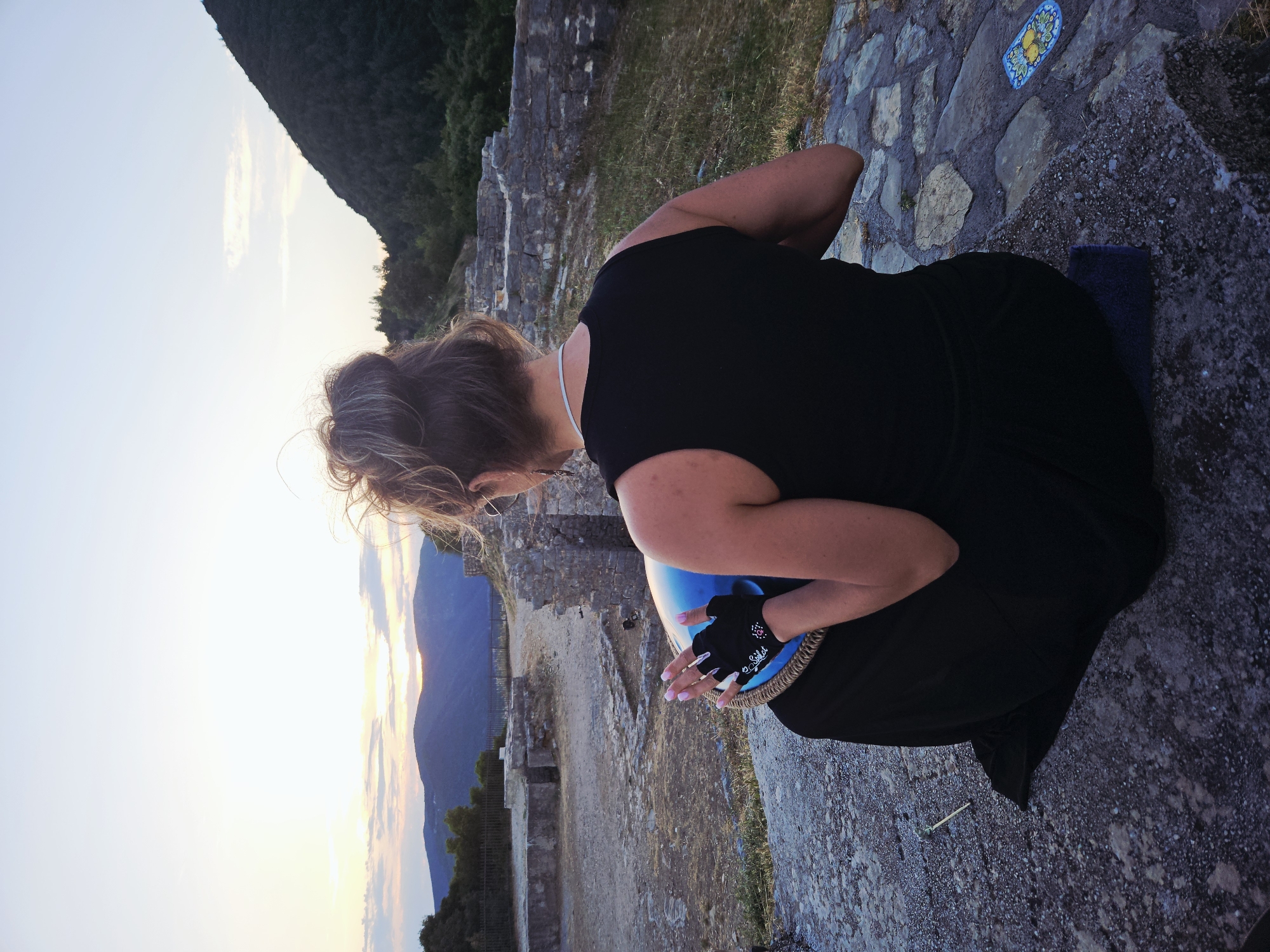 The image, taken from a slightly elevated perspective, shows a woman kneeling or crouching outdoors, viewed from behind and to her right side. She has light brown hair tied up in a bun and wears a black sleeveless top or dress. A thin, light-colored necklace is visible around her neck, along with a small earring. Her hands, clad in black fingerless gloves, hold a vibrant blue, bowl-shaped object with a distinct braided or woven rim. Text "Solos" is visible in white on her left glove.
The scene is set in what appears to be an ancient or historical location, featuring old stone walls and ruins in the midground and background. The ground beneath her is paved with irregular stone slabs, and a small, decorative blue tile with a yellow floral pattern rests on the ground nearby.
Beyond the ruins, a mountainous landscape unfolds under a sky indicative of either sunrise or sunset. The sky displays a soft gradient of warm yellow and orange near the horizon, transitioning to a pale blue overhead, with minimal wispy clouds. The mountains are dark and forested, silhouetted against the glowing sky. The overall ambiance is calm and contemplative, with the woman appearing focused on the object in her hands.