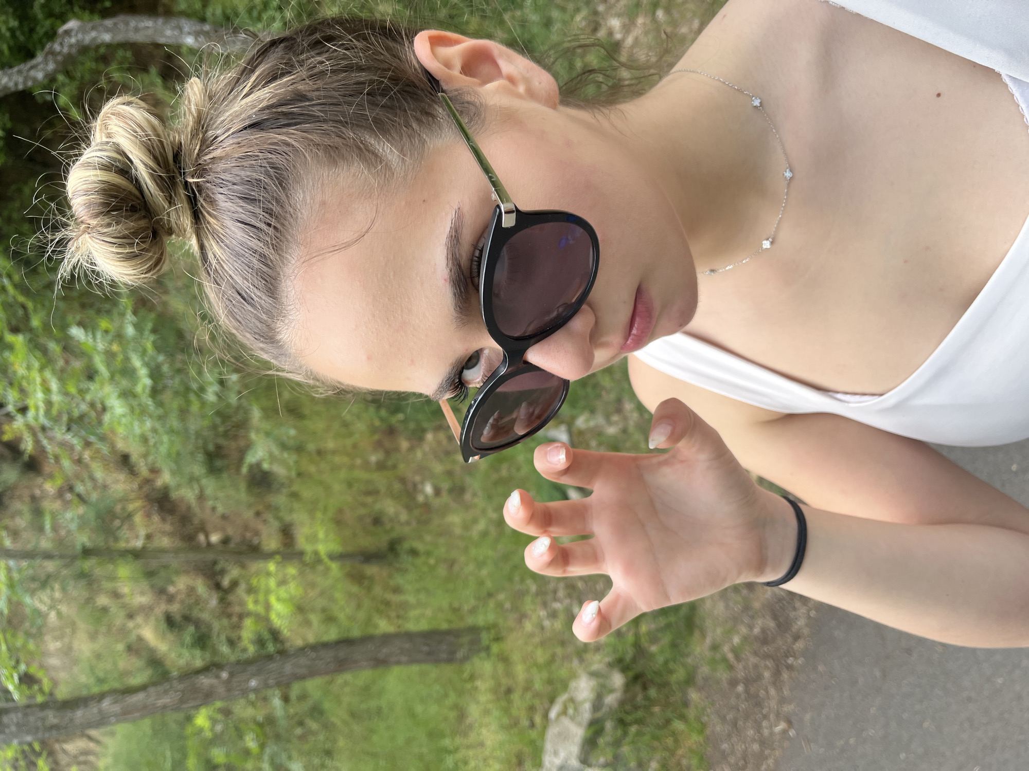 A woman with her hair in a bun is outdoors wearing sunglasses and a white tank top. She has a silver necklace with multiple small pendants. She is wearing a black elastic hair band around her wrist. The background appears to be a wooded area. There is no explicit indication of this being Blagoevgrad, Bulgaria.
