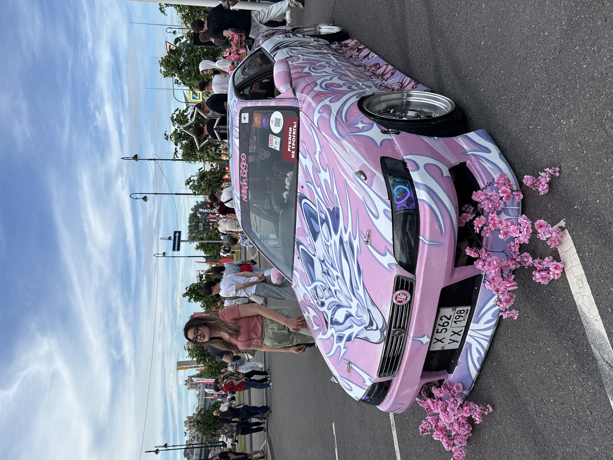 The image captures a custom pink car with a white and purple flame and dragon-like graphic design, parked on asphalt, likely at an outdoor event in Saint Petersburg, Russia. The car features decorative cherry blossom branches draped over its front bumper and a license plate reading "X 562 YX 198 RUS". A young woman with glasses and a pink top is standing next to the car. In the background, a crowd of people, streetlights, trees, and buildings are visible under a partly cloudy sky.