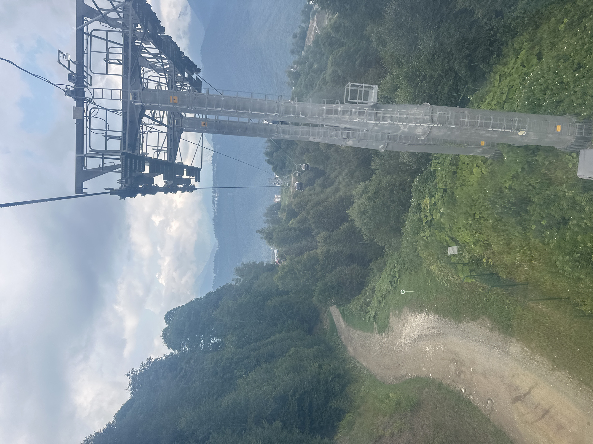 The image shows a view from a ski lift in Estosadok, Russia. The ski lift tower is a prominent structure on the right side of the image, with cables and gondolas visible in the distance. A dirt path winds down a forested mountainside on the left. The sky is overcast with some patches of blue. The vegetation is lush and green, indicating a temperate climate. The scene appears to be a tourist destination, likely in the summer or early autumn, given the greenery and the operational ski lift.