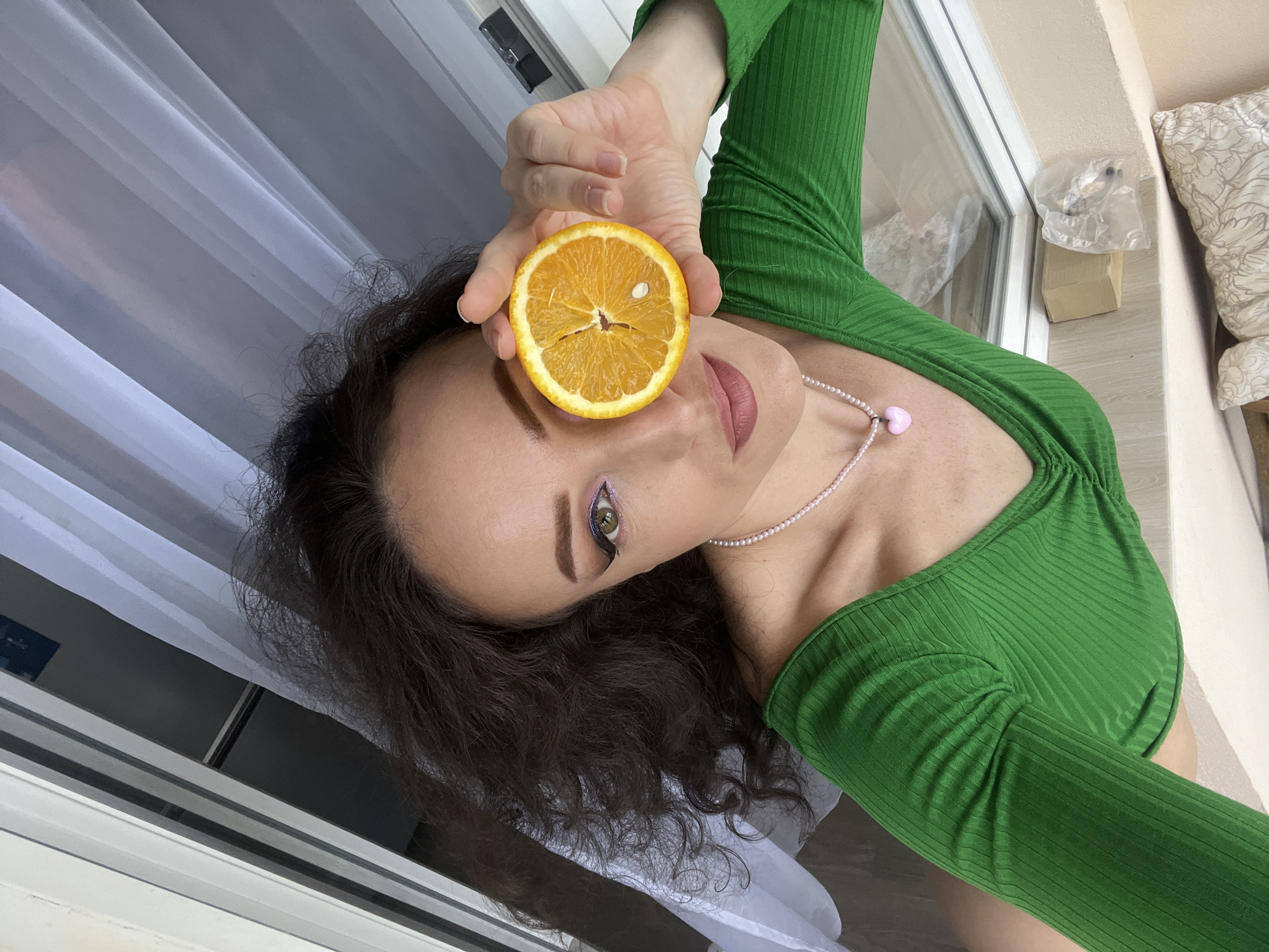 In a high-angle, medium shot, a woman with dark, curly hair is positioned in the center of the frame, holding a slice of orange to her right eye. She is wearing a vibrant green, long-sleeved ribbed top with a V-neckline and a pearl necklace with a pink heart pendant. The background is a white sheer curtain and what appears to be a window frame. The lighting suggests an indoor setting, possibly near a window. The woman's makeup includes dramatic winged eyeliner and pink lipstick. The overall scene is brightly lit, with the focus on the woman and the orange slice. There is no discernible connection to the city of Kazan, Russia, in the image content.