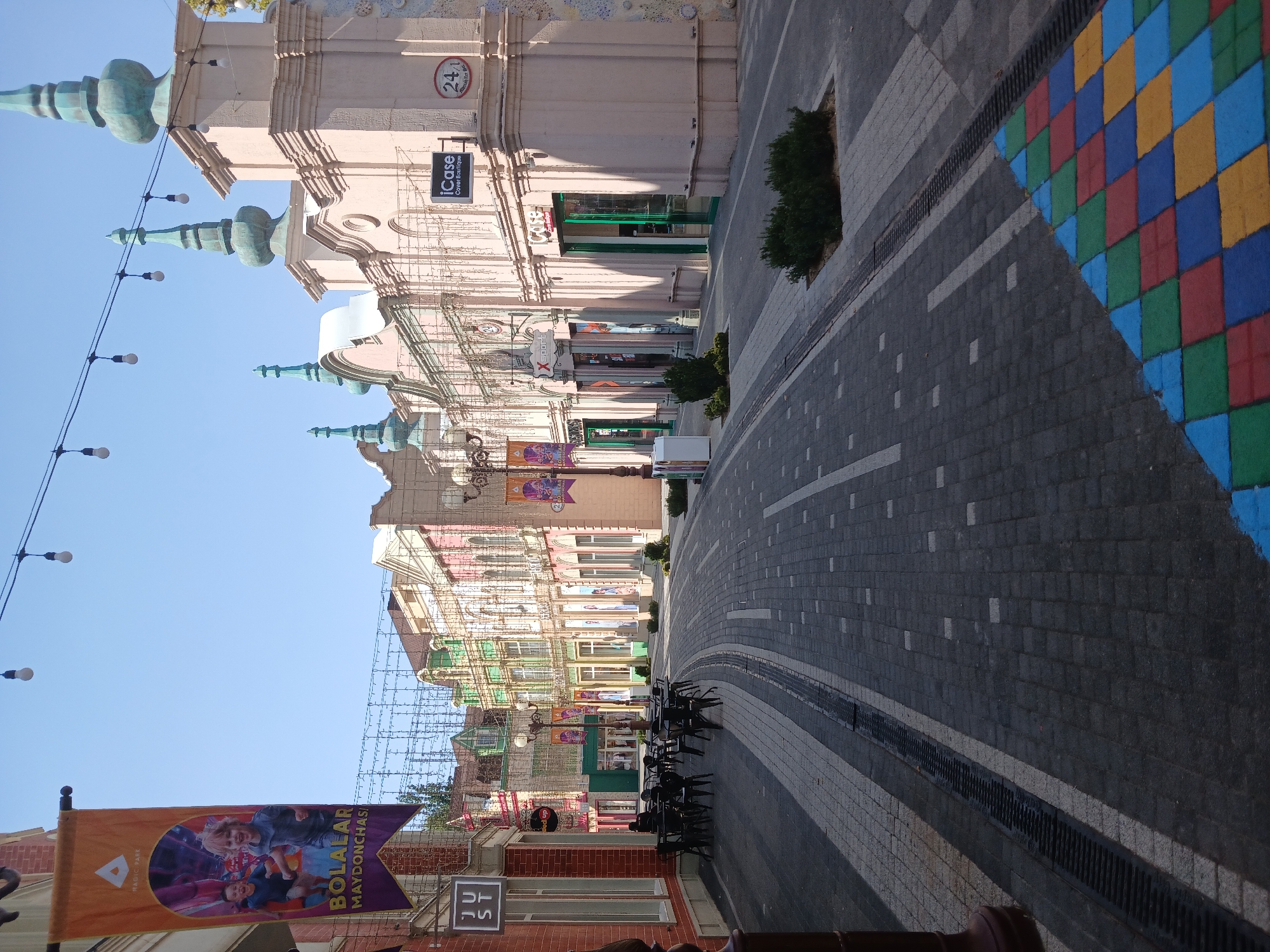 This image depicts a street scene in Tashkent, Uzbekistan, bathed in bright daylight, suggesting a warm and clear day. The perspective is from street level, looking down a gently curving street lined with colorful buildings and a mosaic-tiled walkway. The architecture features ornate facades with pastel colors, decorative columns, and pointed turrets, giving the area a whimsical, almost festive atmosphere.
The street itself is paved with gray cobblestones, interspersed with lighter gray paving stones, creating a patterned effect. On the right side of the street, a section of the walkway is adorned with a vibrant mosaic of colorful squares in red, blue, yellow, and green, resembling a checkerboard pattern. This colorful detail adds a playful element to the urban landscape.
Several buildings are visible, with shop fronts and signage. One banner, prominently displayed on the left, features an image of children playing and text that reads "BOLALAR MAYDONCHAS" and "MAGIC PARK," indicating a children's play area or amusement park nearby. Other signs are visible but not clearly legible.
Objects present include streetlights, strung with decorative bulbs, casting shadows on the buildings and walkway. Dark-colored chairs are neatly arranged in front of some of the establishments, suggesting an outdoor seating area for cafes or restaurants. A few small, well-maintained green bushes are planted along the street. The overall impression is that of a well-kept, lively public space, likely a commercial or entertainment district. The scene is quiet, with no people visible, implying it might be early in the day or during a less busy period. The clear blue sky suggests good weather.