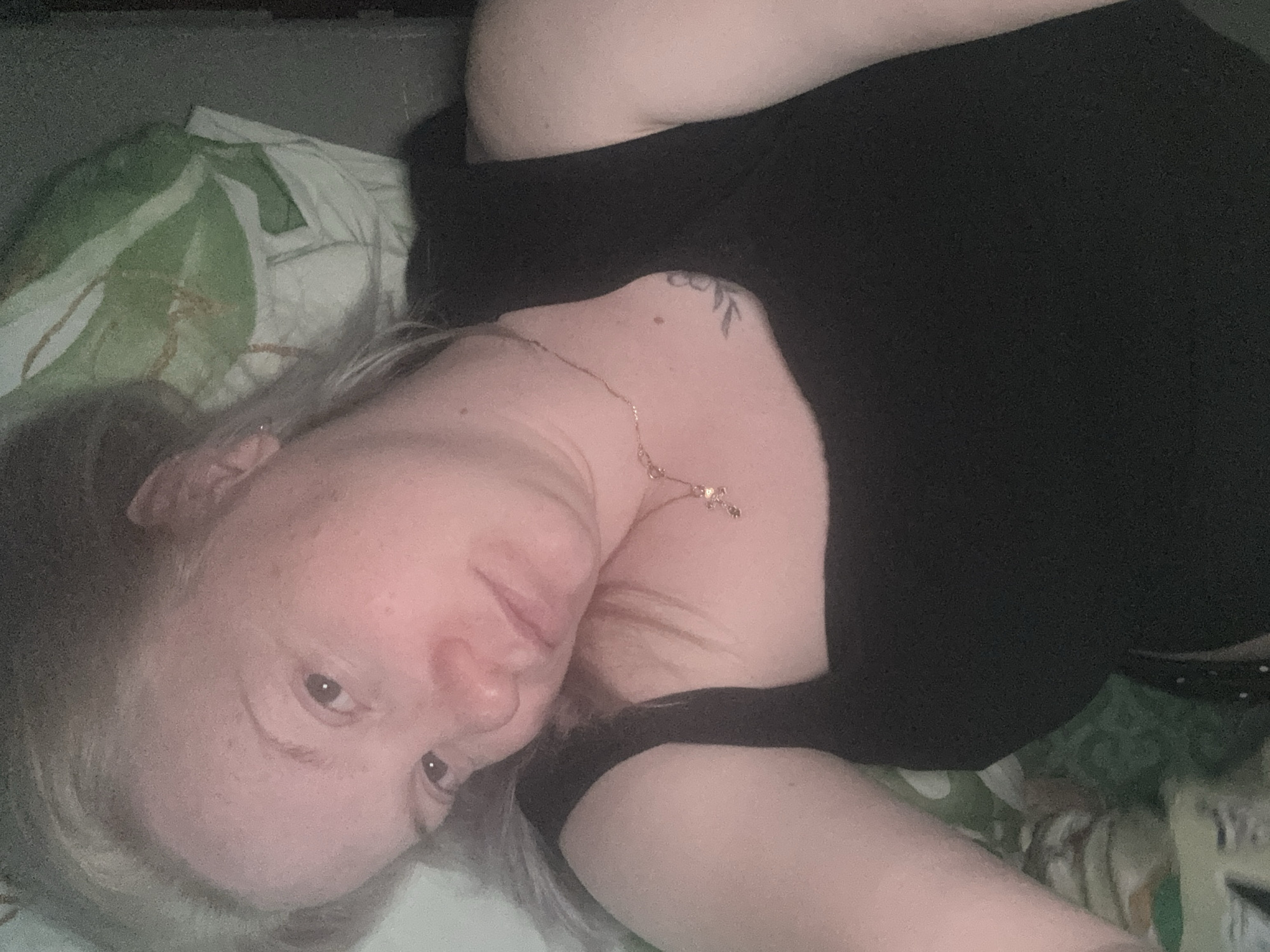 The image captures a close-up, slightly angled indoor portrait of a person, likely a selfie. The subject is an adult with fair skin and light-colored hair, possibly blonde or light brown, and appears to be lying down, facing the camera with a neutral and relaxed expression.
The person is wearing a black garment, such as a t-shirt or top. Around their neck is a delicate gold-colored necklace with a small, indistinct charm, possibly a cross. A noticeable tattoo, depicting a leafy branch design, is visible on their upper back or shoulder area.
The background features patterned bedding or a pillow, displaying a green and white abstract or floral design. A dark, solid surface, potentially a headboard or wall, is visible behind the bedding. The lighting is dim and soft, suggesting an indoor setting during the evening or night. The overall atmosphere is calm and personal, indicative of a private domestic space, potentially a bedroom. No text is visible in the image. The setting is consistent with a home environment in Chaikovsky, Russia.