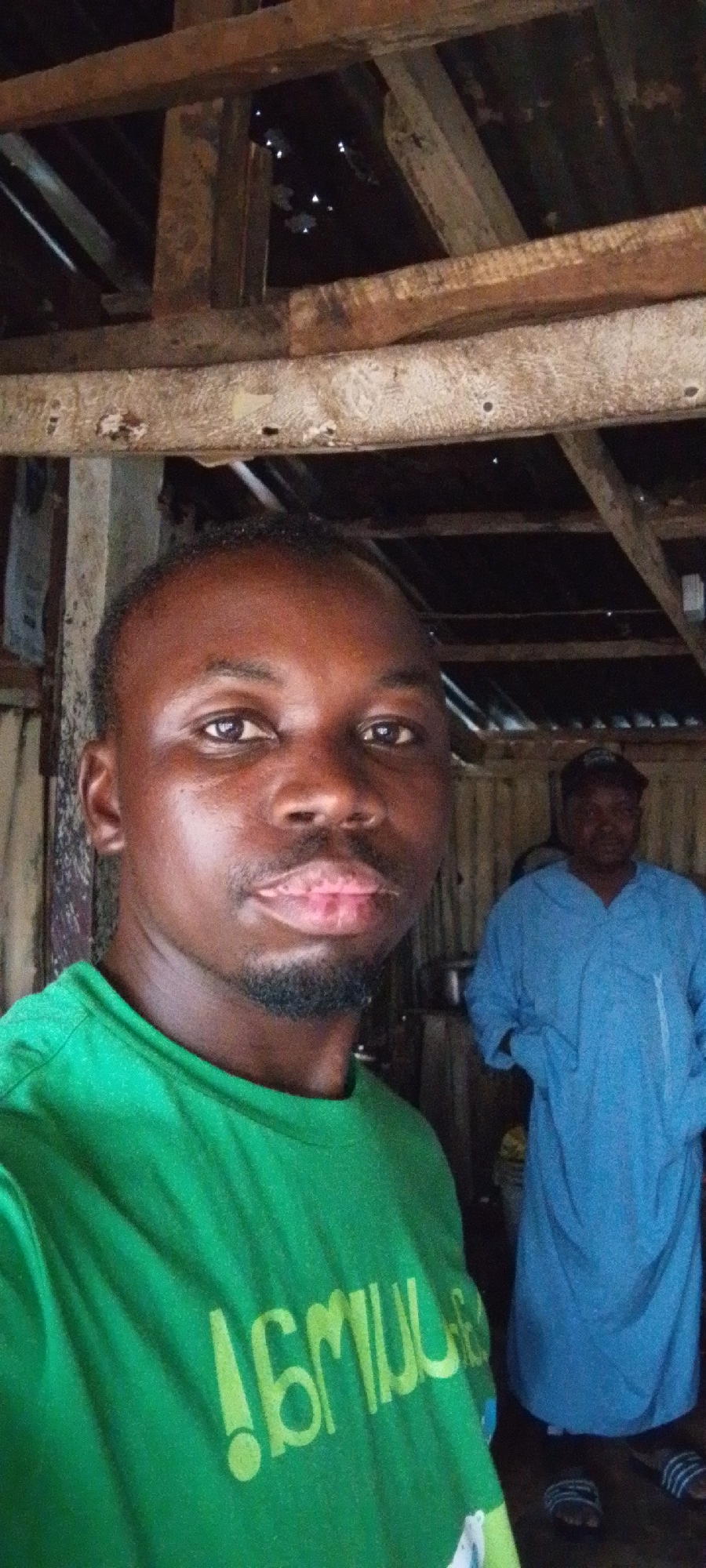 A man wearing a green t-shirt is taking a selfie. Behind him, another man wearing a blue robe and slippers stands in a room with exposed wooden beams and a corrugated metal roof. Based on the clues in the image and context of the city of Jos, Nigeria, the location appears to be in a modest building or home possibly in a rural part of Jos.