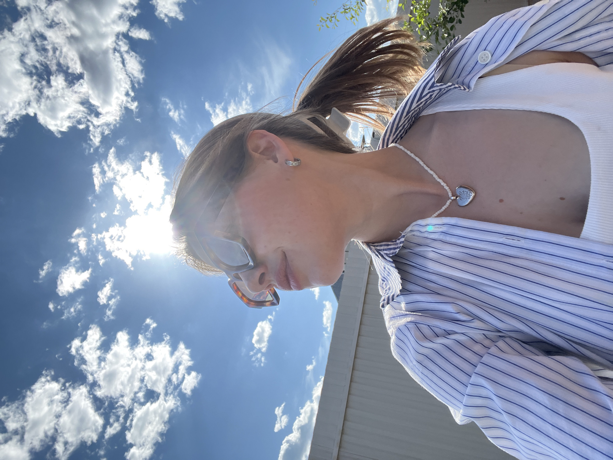 The image depicts a young adult female from the upper chest up, positioned slightly to the right of the frame, looking towards the upper left. She is wearing large, tortoise-shell framed sunglasses, a white ribbed tank top, and an open, light blue and white vertically striped button-down shirt with the sleeves pushed up. A white beaded necklace with a silver heart-shaped locket is visible around her neck. Her light brown hair is pulled back into a low ponytail, secured with a white claw clip, and a small silver hoop earring is present in her visible right ear. She exhibits a subtle smile.
The scene is outdoors under a bright, clear blue sky with scattered white cumulus clouds. Direct sunlight from the upper right creates a prominent lens flare and strong highlights across the subject and the sky. To the left of the subject, a light-colored, possibly off-white or light grey, corrugated metal fence or wall extends horizontally. On the far right, some green foliage from a bush or tree is visible. This outdoor setting is presented within the context of the city of Chaikovsky, Russia.