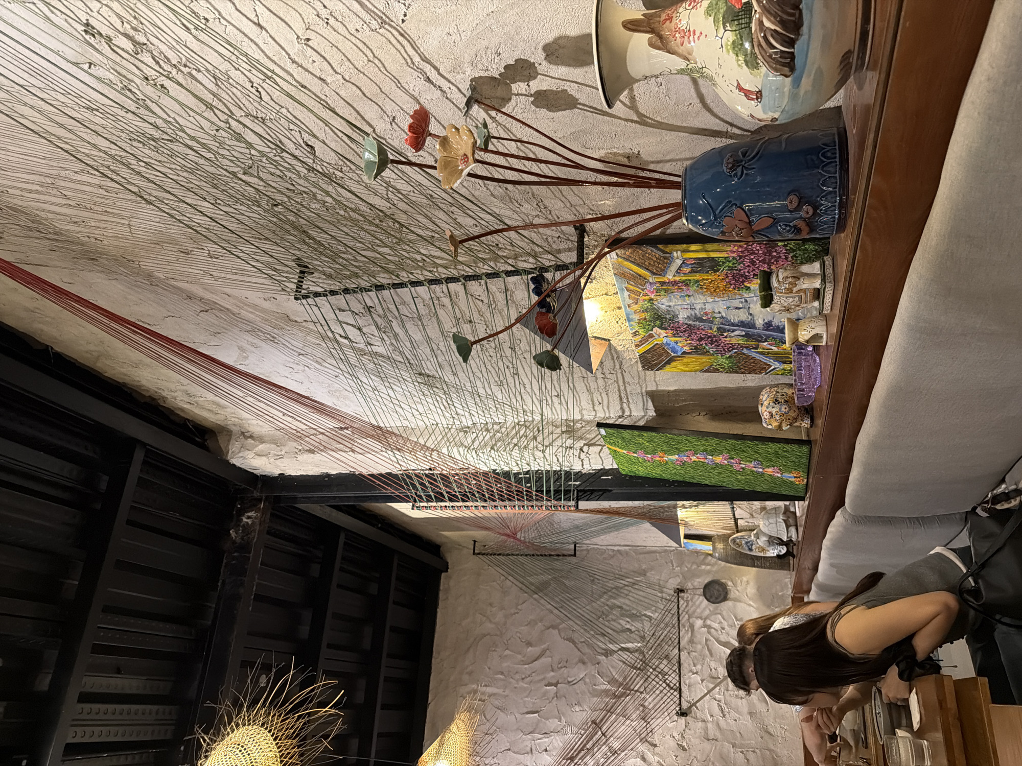 The image shows an interior space in Đà Nẵng, Vietnam. The ceiling is decorated with numerous hanging strings of various colors, mainly green and red, creating an artistic installation. Beneath the ceiling are black metal beams and a textured white wall. Part of the ceiling is composed of dark horizontal slats, contrasting with the light-colored areas.
On the right side of the image, there is a shelf displaying decorative items, including a ceramic vase, a blue vase with floral designs, and a framed picture. Part of a gray sofa is also visible. Two people are present near a wooden counter, appearing to be dining or engaging in an activity at the counter. One person has long dark hair, and part of another person is also seen.