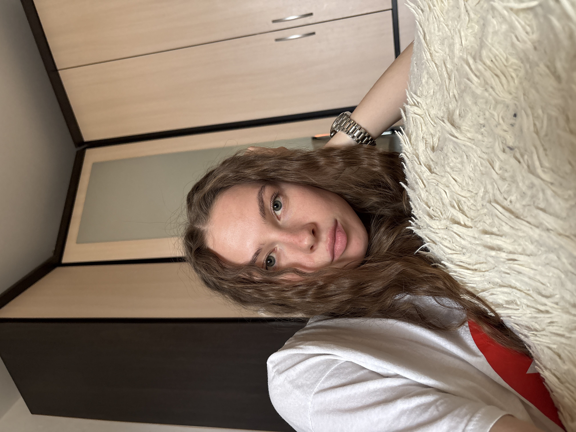 A young woman with long, curly brown hair and light green eyes is lying down, resting her head on a fluffy, cream-colored throw pillow. She is wearing a white t-shirt with a red design on the left side. Her left arm is bent, and she is wearing a silver watch on her wrist. She is looking directly at the camera with a neutral expression. Behind her, there is a closet with two doors, one dark brown and one light wood-colored. The lighting suggests it is daytime. The environment appears to be a bedroom or a similar domestic setting. The image does not contain any information that would allow for determining a specific location in Perm, Russia.