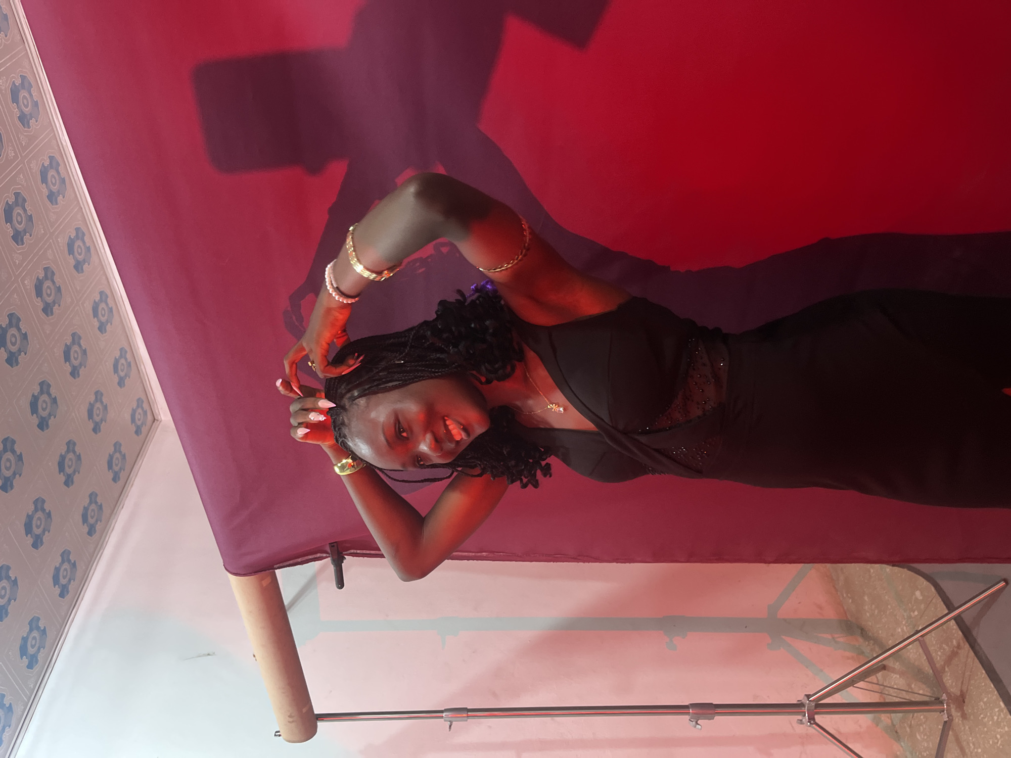 The image depicts a young Black woman posing in what appears to be a makeshift photo studio. She is standing in front of a deep red backdrop, with a rolled-up burgundy fabric visible to her left, supported by a lighting stand. The ceiling features a white patterned design. The woman is wearing a black dress with a deep V-neck and embellished detailing around the bust. She has long, dark braided hair and is wearing multiple gold bracelets on her right arm, along with a pearl bracelet on her left wrist. Her fingernails are long and painted white. She is smiling and has her hands behind her head, with her tongue sticking out playfully. A shadow of the woman is cast on the red backdrop behind her. The lighting appears to be artificial, with a red hue casting over the scene. The floor is concrete. There are no specific indicators within the image that definitively place this scene in Gura Top, Nigeria, or any other specific city.