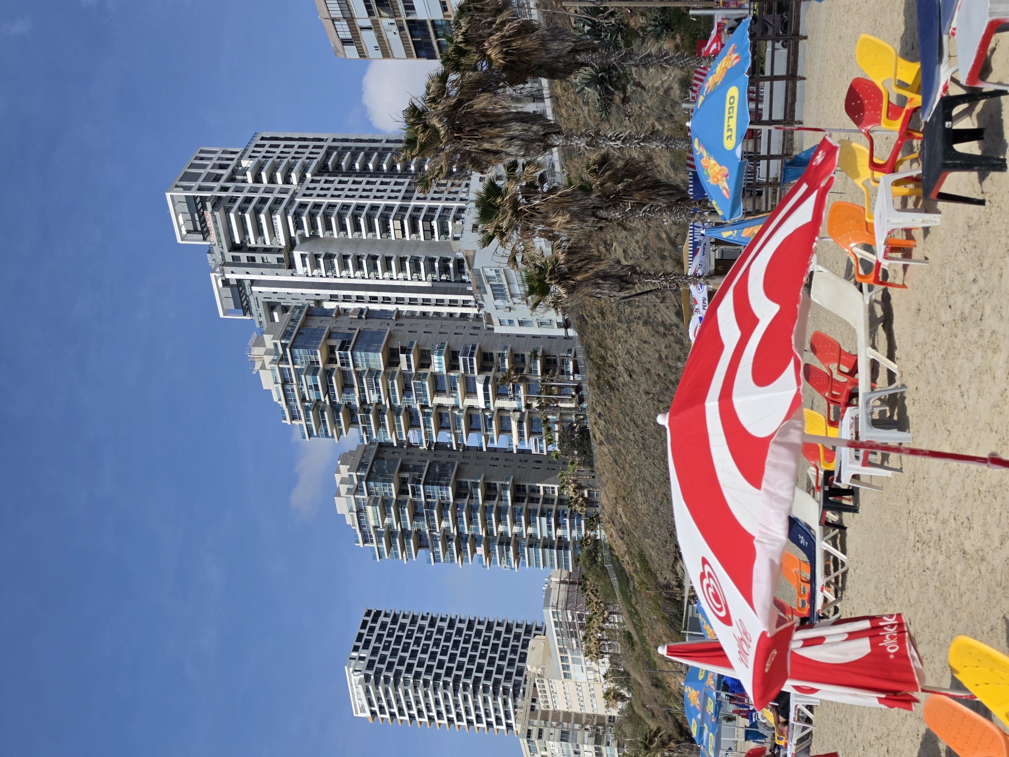 This image, taken in Bat Yam, Israel, depicts a beach scene with high-rise buildings in the background. The blue sky is visible above the buildings. The beach has colorful umbrellas and chairs. No people are visible in the image.