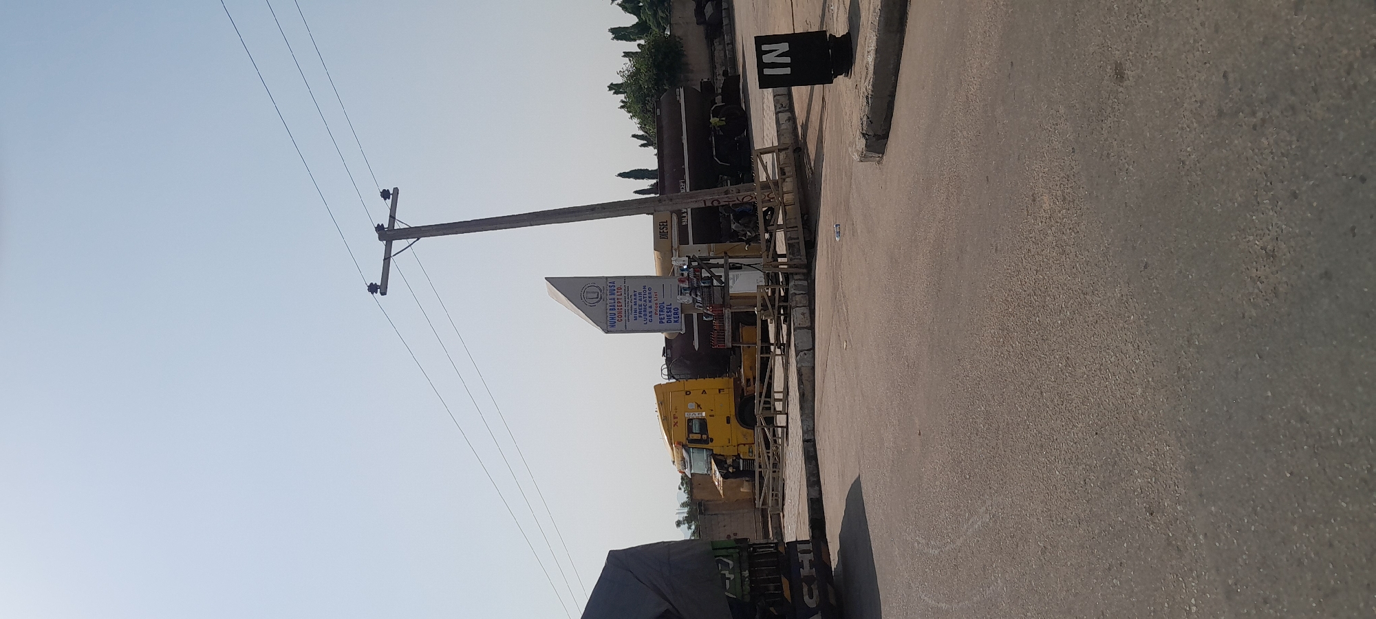 In Jalingo, Nigeria, a daytime scene unfolds at a gas station. A yellow semi-truck, bearing the "XF" insignia, is parked near a gas pump. Adjacent to it, a long, dark tanker truck is also positioned. A white sign with a blue logo and text reads "NUHU BALA MUSA CONCEPT LTD." below which is a price list for PETROL, DIESEL, and KERO. A black sign with white lettering at the forefront reads "IN," indicating an entrance. The sky is a clear, pale blue. Electrical poles and wires are visible, contributing to the urban setting. The ground is paved, suggesting a road or parking area.