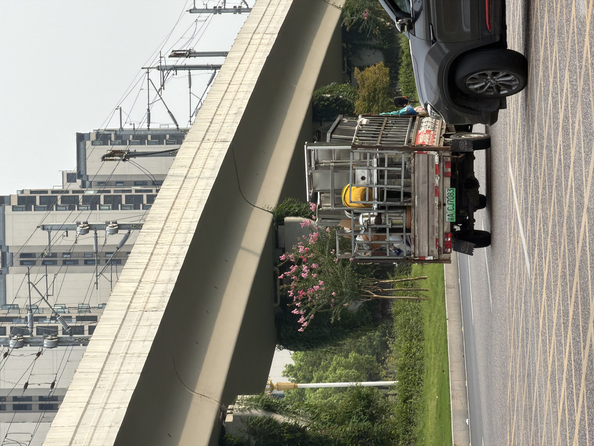 In Jiangjin, China, a gray SUV is traveling on a multi-lane road. The SUV is in the right lane, partially obscuring a flatbed truck in the adjacent lane. The truck is carrying a load of metal shelving and a yellow barrel. The truck's license plate reads "A CJ7083". In the background, elevated train tracks supported by concrete pillars run parallel to the road, with buildings and trees visible beyond. The sky is overcast.