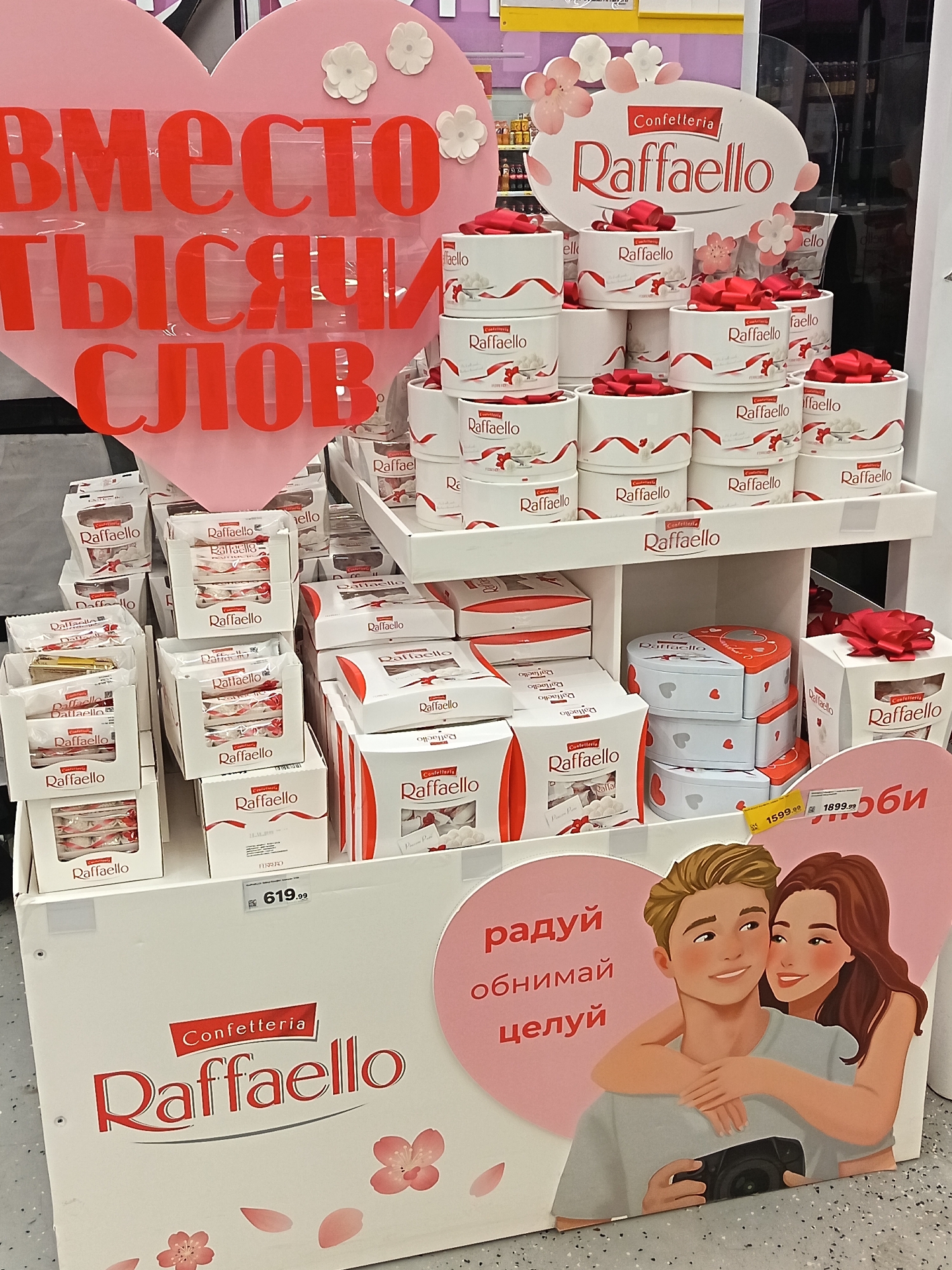 The image shows a retail display of Raffaello candies in a store, likely in Nizhnevartovsk, Russia based on the Cyrillic text present. The display is primarily white, with red and pink accents.
The candies are stacked in various forms, including round boxes with red ribbons, rectangular boxes, and wrapped candies. There are promotional materials with the Raffaello logo and text in Russian. Some text includes "Вместо тысячи слов," translating to "Instead of a thousand words," and "радуй обнимай целуй" which translates to "rejoice, hug, kiss." There is also an image of a couple hugging. Price tags are visible, with prices such as 619.99, 1599.99, and 1899.99. The overall setting appears to be a supermarket aisle.