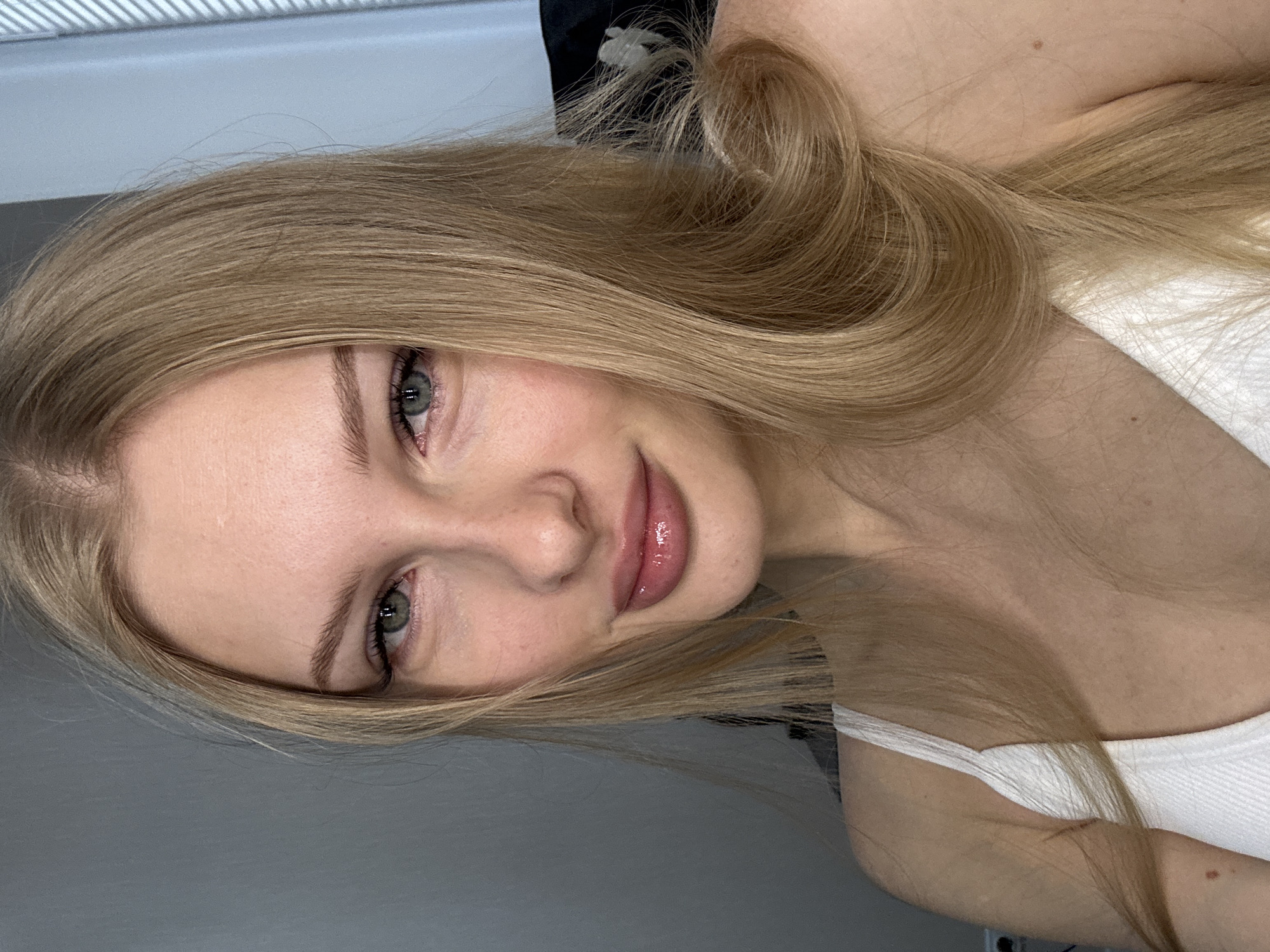 The image is a close-up, head and shoulders shot of a young Caucasian woman with blonde hair and green eyes. She is wearing a white ribbed tank top. Her makeup is subtle, with a focus on her lips, which are glossy and a pinkish-nude shade. Her hair is parted to the side and falls over her shoulders. The background is indistinct, with a hint of a window blind on the right side and a dark object behind her on the left. The lighting is soft and even, with no harsh shadows. The overall impression is one of natural beauty and relaxed confidence.
The provided image is a portrait and does not contain any elements that would allow for the identification of its location in the city of Perm, Russia, or any other specific geographic location.