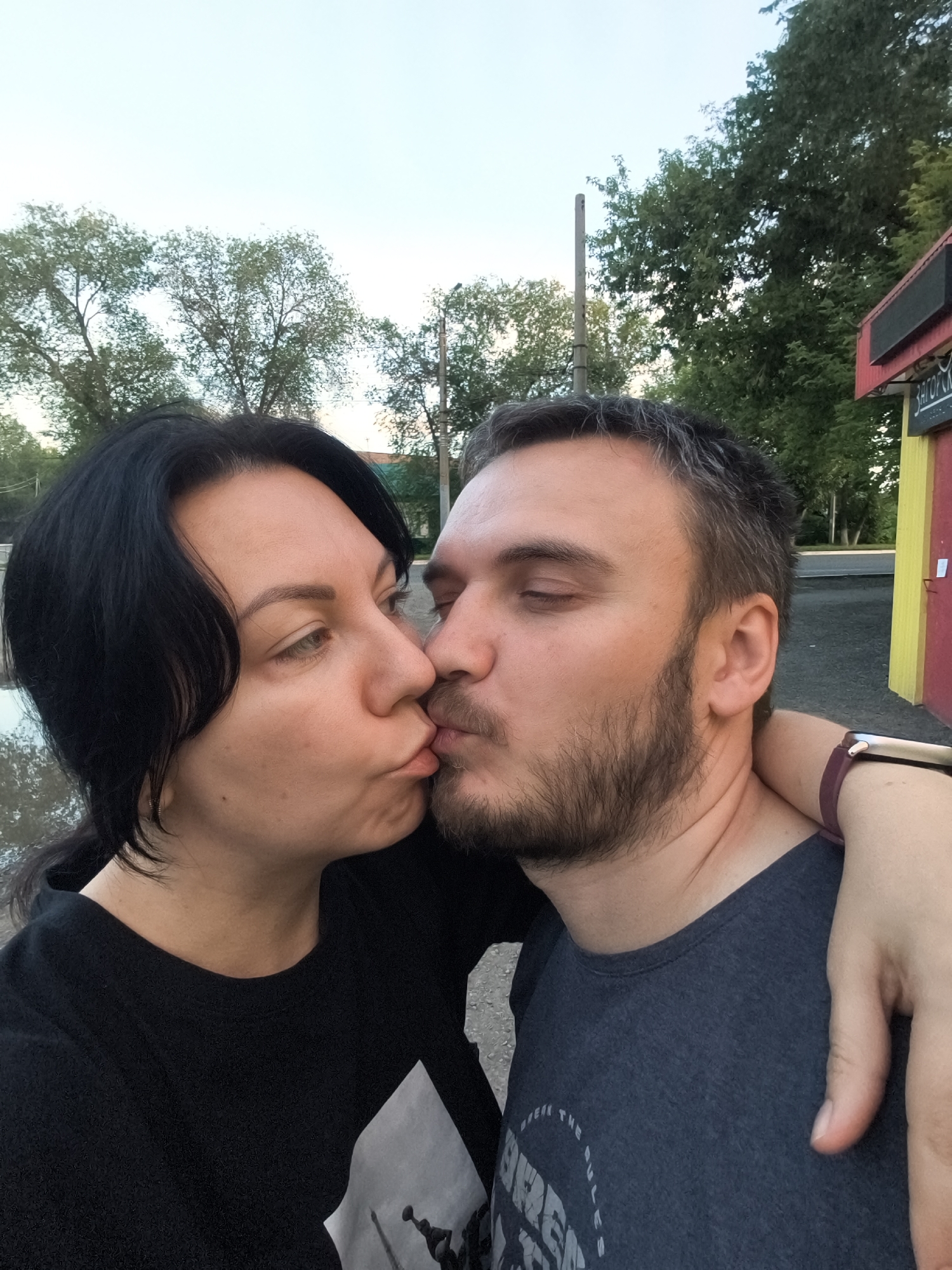 The image shows a couple kissing outdoors, with trees and a building visible in the background. The woman, on the left, has dark hair and is wearing a black t-shirt. The man, on the right, has short dark hair with graying temples and a beard, wearing a dark grey t-shirt. His eyes are closed. The woman's arm is around the man's neck. The scene appears to be a street or park area. The building in the background is red and yellow with some signage. Based on the architectural style and vegetation, the location is consistent with an urban or suburban setting in Orenburg, Russia.