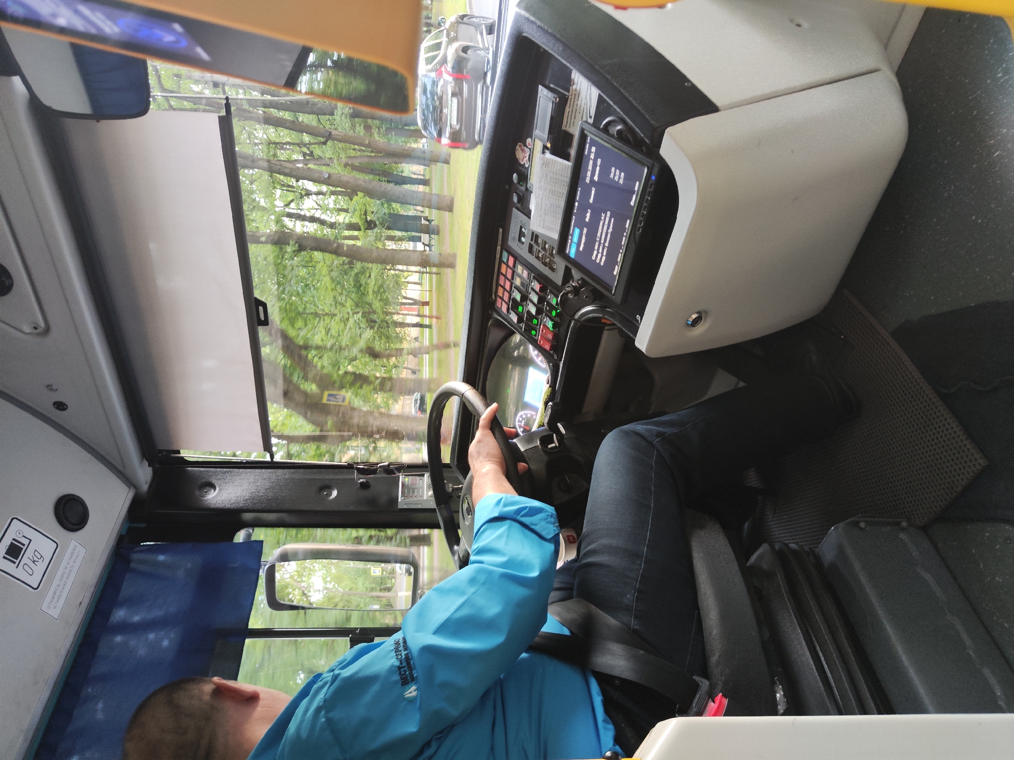 This media file captures the interior of a bus from the perspective of the driver. A person wearing a bright blue jacket and dark jeans is seated in the driver's seat, gripping the steering wheel. The driver's face is partially visible, showing the back of their head and ear. The bus's dashboard is prominent, featuring a digital screen displaying information that includes "20.05.2022" and "22:55:00", suggesting the time and date. Various buttons and controls are visible on the dashboard. Outside the bus, trees and greenery are visible through the windows, indicating an outdoor setting. The lighting suggests it is daytime, possibly late afternoon or early evening due to the time displayed on the screen. The scene appears to be within the city of Pushkin, Russia, as indicated by the location context. There is a caution sign on the left side of the bus interior with text that includes "0 kg".
