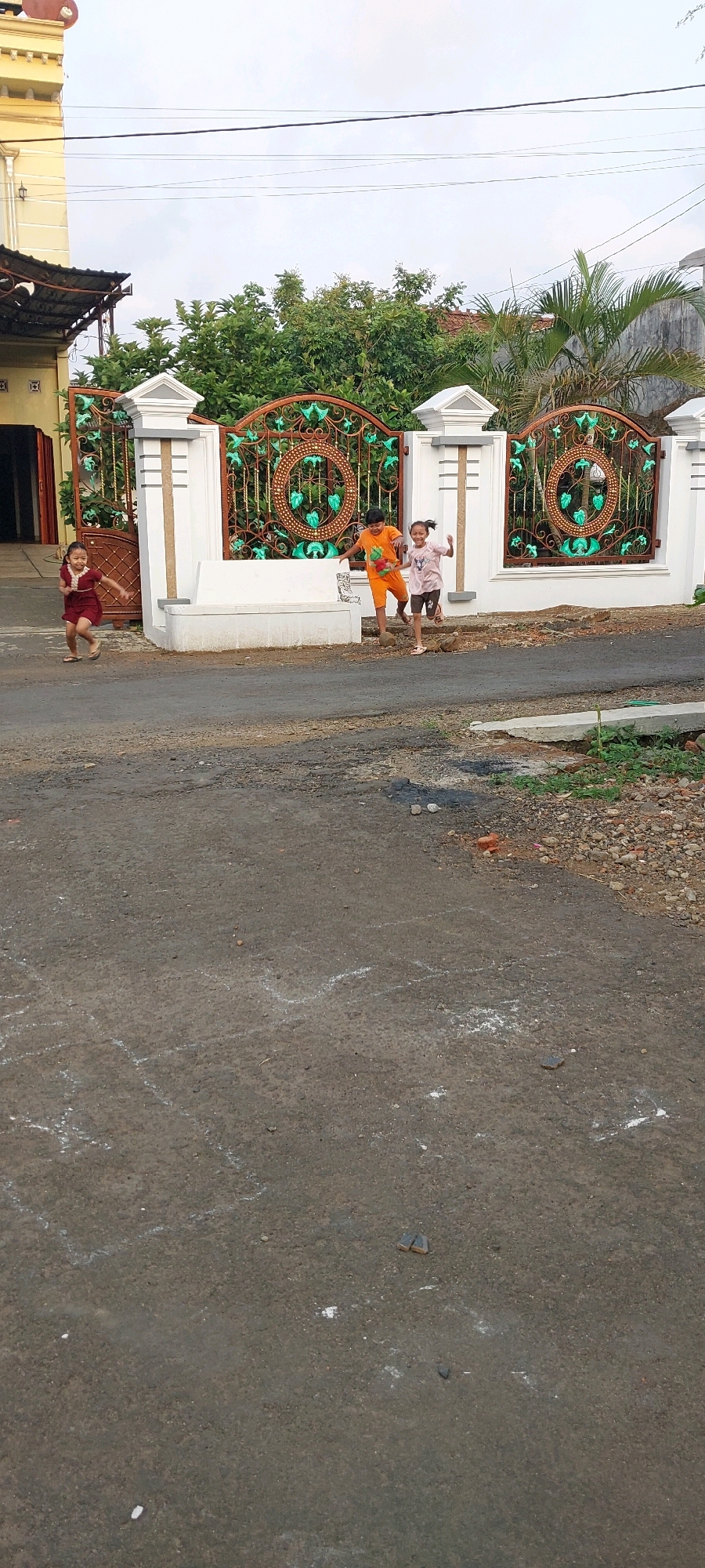 In the city of Keling, Indonesia, three children are captured in motion on a paved road in front of a white and brown ornate gate. The child on the far left, a girl in a maroon dress, is running towards the left, her arms extended. To her right, a boy in an orange shirt and shorts is mid-stride, appearing to run towards the viewer while looking to his left. Next to him, another child, possibly a girl in a pink shirt and brown shorts, is running towards the right with their arms outstretched. The gate behind them features decorative circular patterns with green accents. The road surface is asphalt, with white chalk markings visible, suggesting a hopscotch game. The environment includes a building to the left, lush green trees and foliage behind the gate, and a slightly overcast sky.
