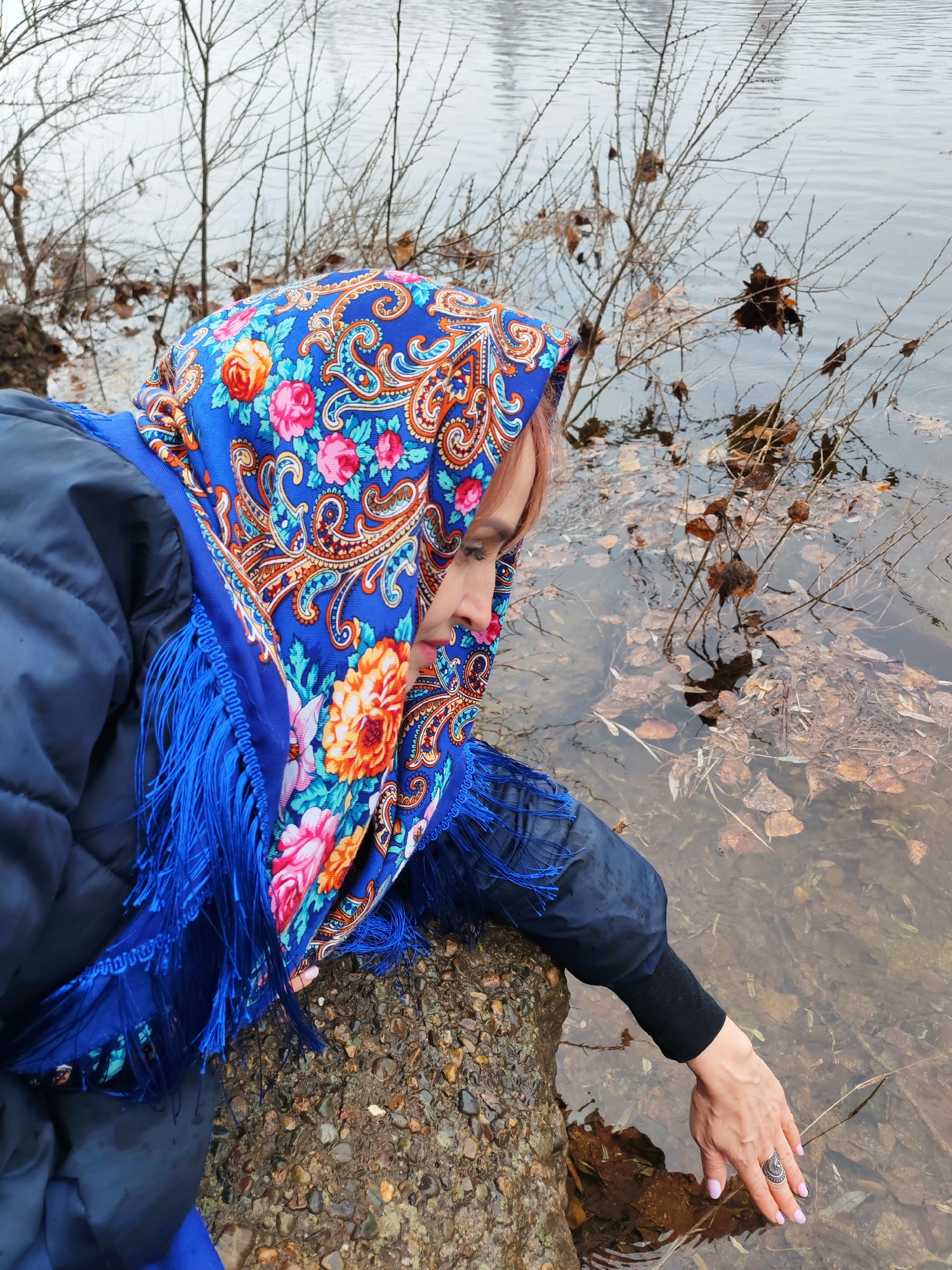 A woman with red hair is kneeling by the edge of a body of water, wearing a blue floral headscarf with fringe and a dark blue quilted jacket. Her right hand reaches into the water, which is filled with fallen leaves and small branches. A stone bank is visible beneath her. Leafless bushes line the water's edge in the background, and the body of water stretches out into the distance under an overcast sky. The scene is in Chaikovsky, Russia.
