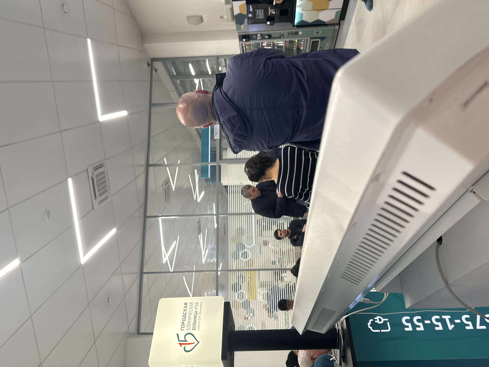 The image shows an interior shot in what appears to be a medical facility or clinic in Moscow, Russia. Several people are present, including individuals in a queue. The environment is a modern, well-lit space with a white tiled ceiling. A sign in Russian is visible that reads, in part, "Городская Клиническая Больница", indicating a city clinical hospital. The text "575-15-55" is visible, indicating a phone number. A self-service kiosk dominates the foreground.