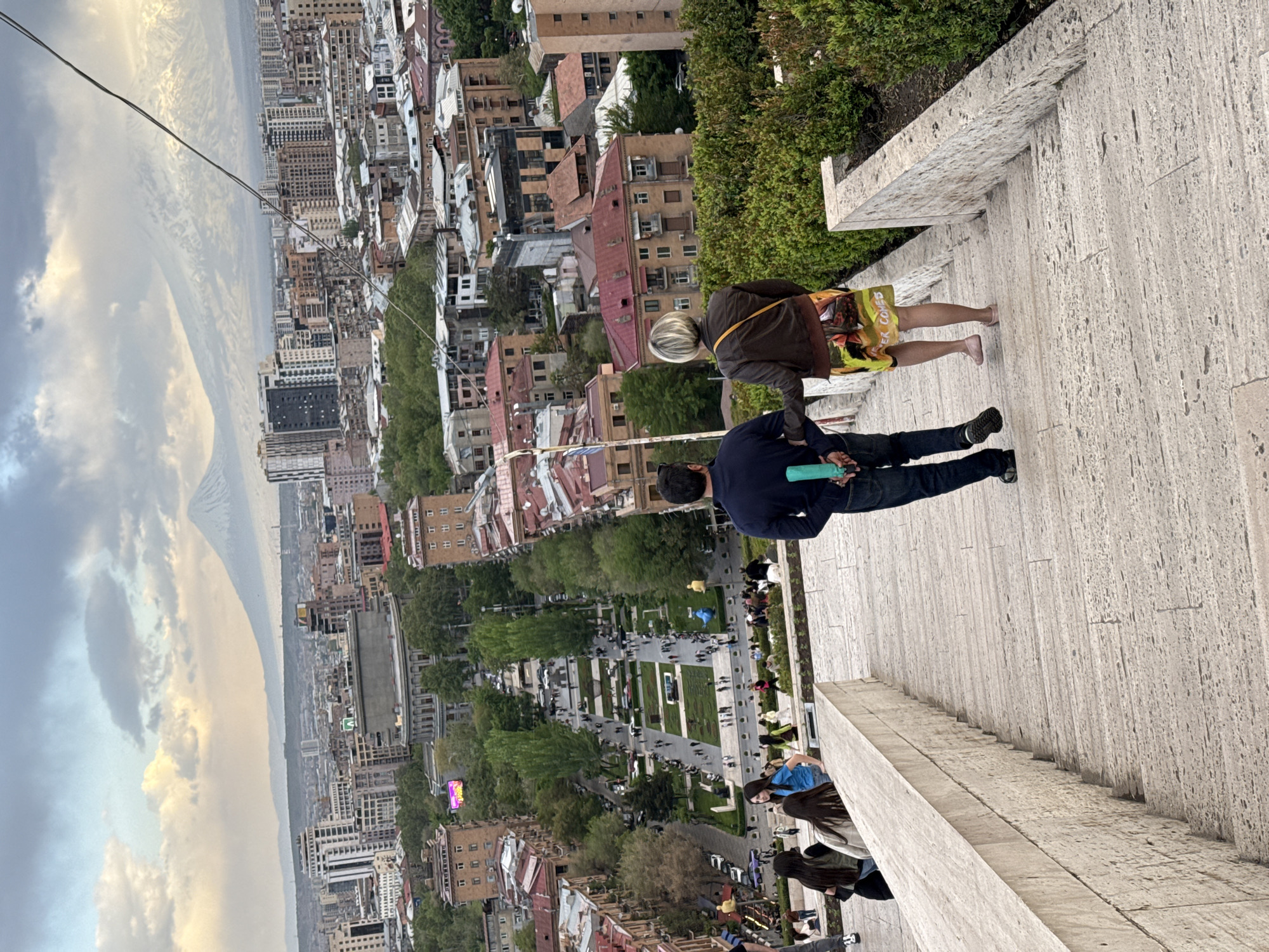 The image captures a high-angle view of the city of Yerevan, Armenia, likely taken from the Yerevan Cascade. In the foreground, two people are visible on the stone steps: one in a dark jacket and patterned shorts, and another with a navy shirt and jeans, holding a green bottle. The steps are large and made of light-colored stone, lined with greenery. Below the steps, a garden area with trees and paths can be seen, with people strolling around. In the background, there's a densely built urban landscape with buildings of varying heights and colors, mostly with red-tiled roofs. Further in the distance, a mountain range can be observed under a cloudy sky. A cable is suspended across the top left of the frame.