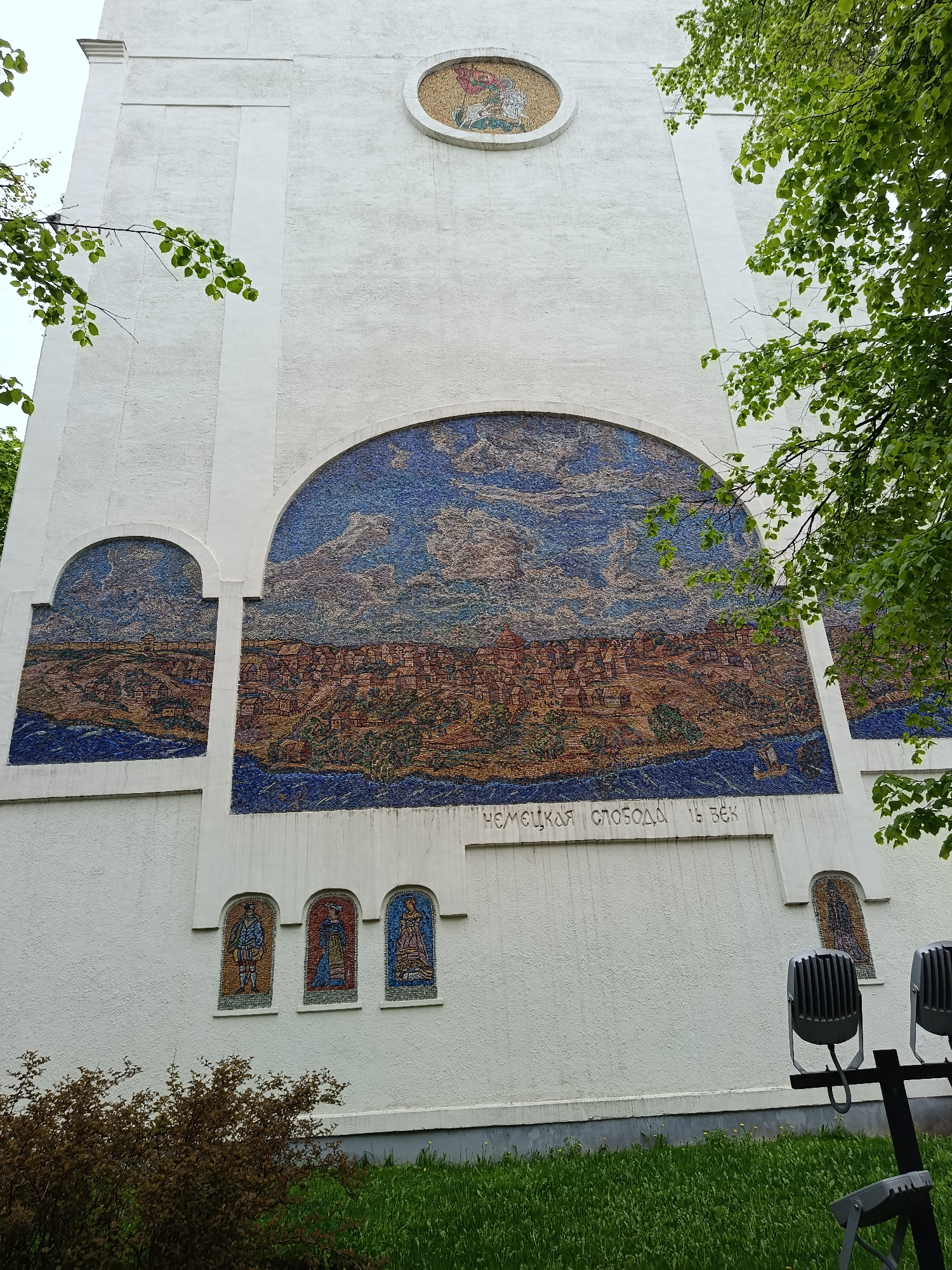 The image shows a large, white building facade adorned with several mosaic murals. The largest mural, an arched mosaic, depicts a cityscape with a river in the foreground and a cloudy sky above. To the left of this large mural is a smaller, similarly arched mosaic showing a related scene. Above the large mural is a circular mosaic, possibly depicting religious figures. Below the large mural, there are three smaller, arched mosaic panels showing individual figures. To the right of the large mural, another similar mosaic panel is visible. Inscribed below the main mosaic in Russian text are the words "Немецкая слобода 16 век" (German Quarter 16th century). This refers to the historical German Quarter of Moscow.
At the bottom of the image, there is grass and some shrubbery. On the right side of the image, a lighting fixture with two spotlights is visible. The perspective of the image suggests it was taken from ground level, looking up at the building. Tree branches with green leaves frame the building on both sides. The building's texture appears to be rough or stucco-like.